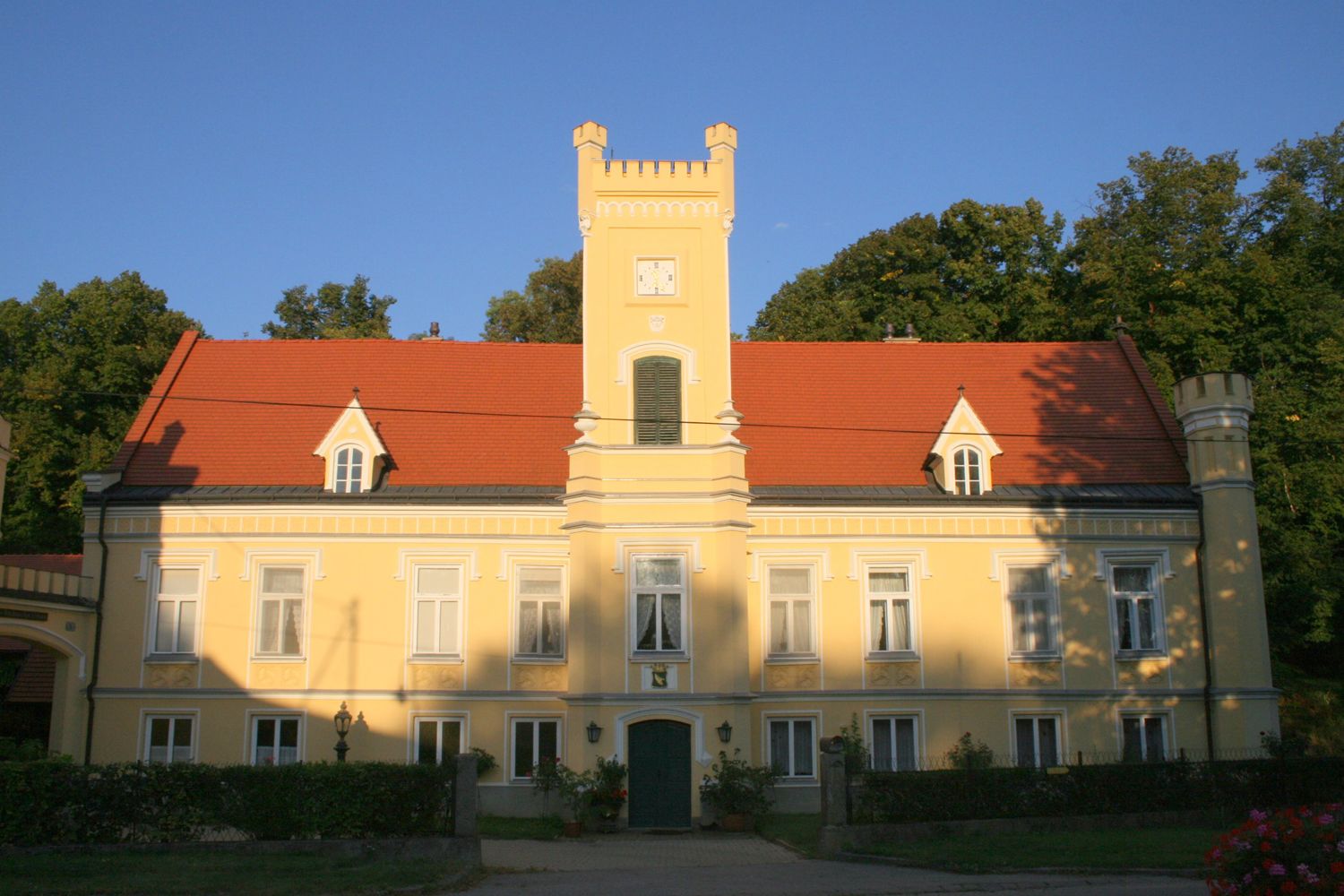 Yellow castle with red roof and tower, surrounded by trees.