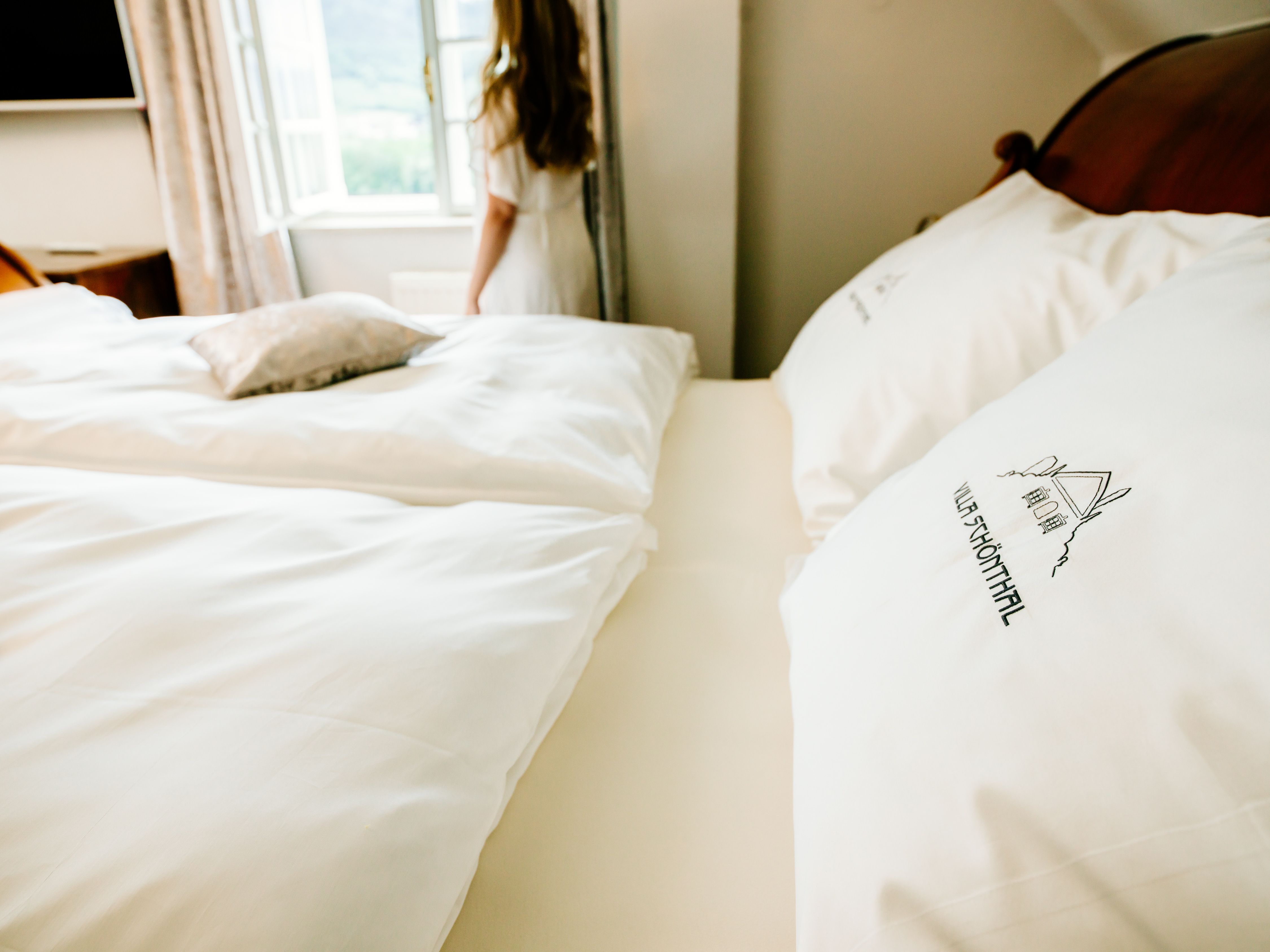 A bed with white pillows and bed linen with "Villa Schönthal" written on it. A person stands at the window and looks out.