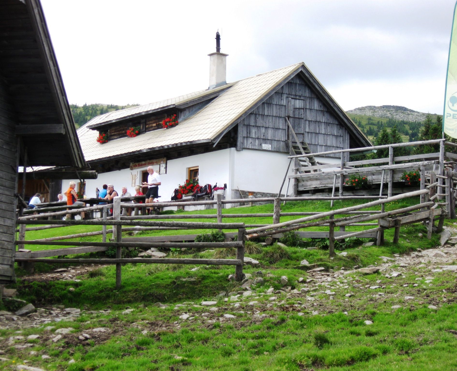 Alpine hut with people in a meadow, surrounded by wooden fences.