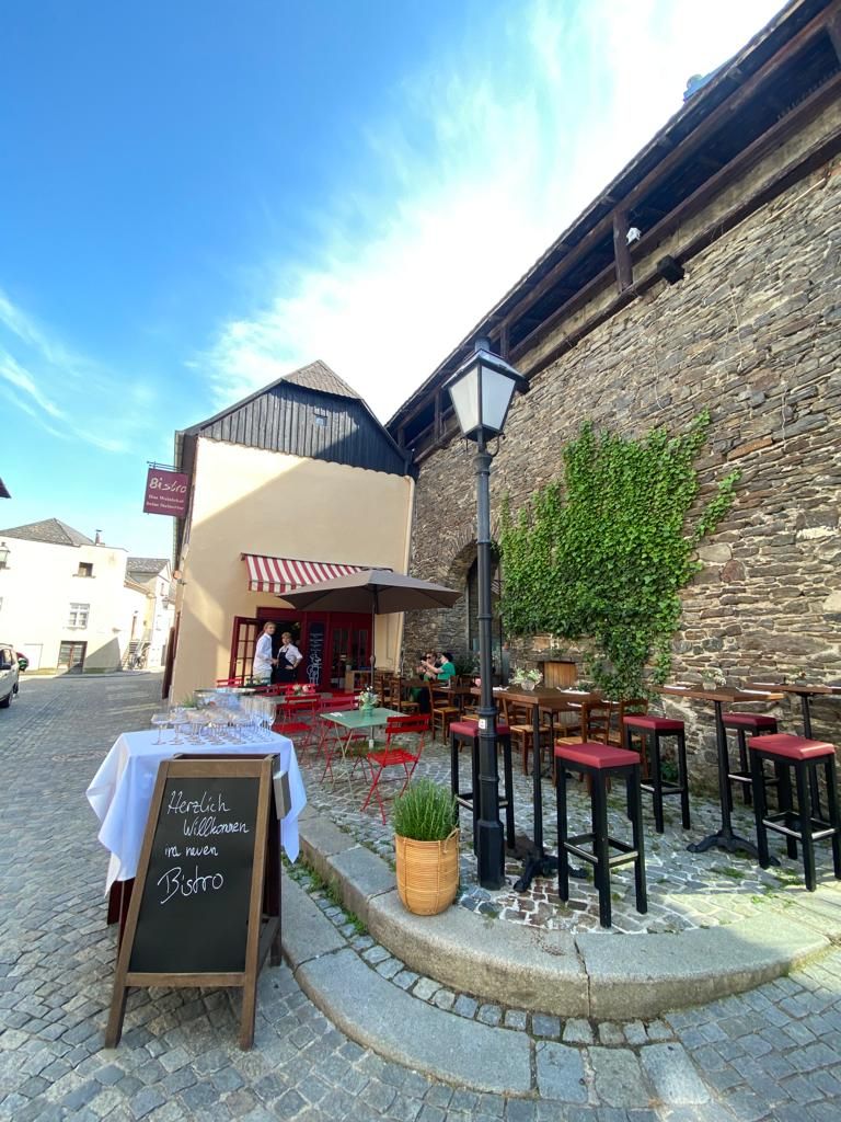 A cozy bistro with red chairs and outdoor tables, surrounded by old stone walls and plants.