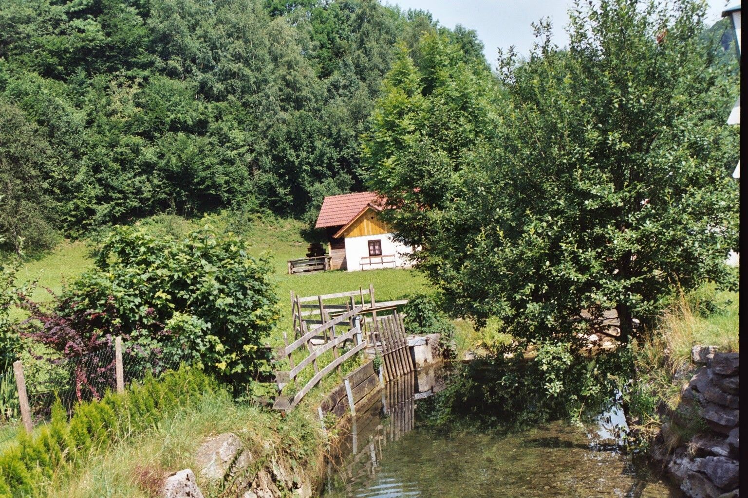 A small hut with a red roof stands in a green landscape next to a stream and trees.