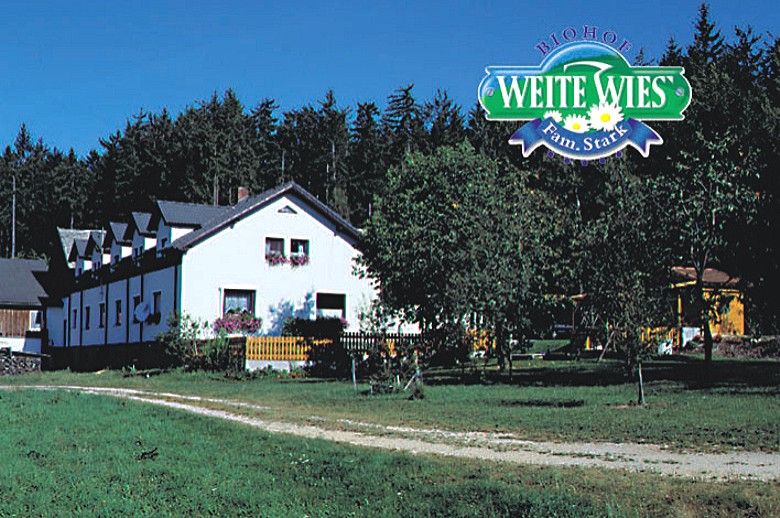 A white farmhouse with flowers in front of a forest, above it the logo 'Biohof Weite Wies Fam.