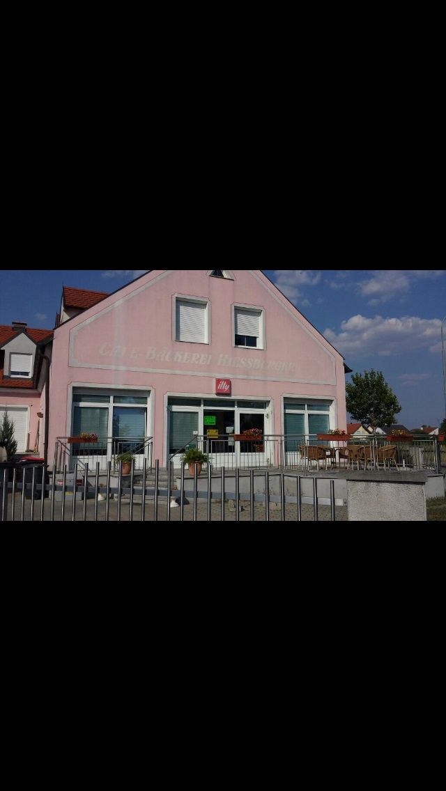 Exterior view of a pink building with the inscription 'Bäckerei-Café Hiessberger'. There are tables and chairs in front of the building.