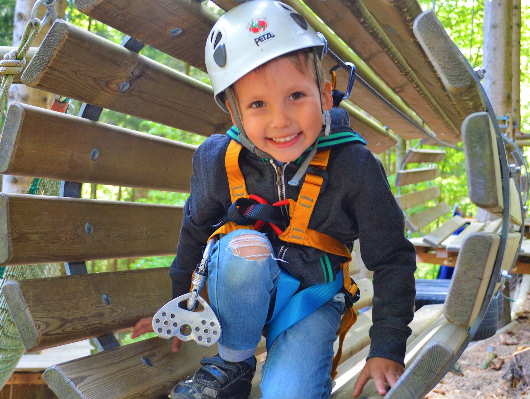 A smiling child with a helmet and climbing harness on a wooden course.