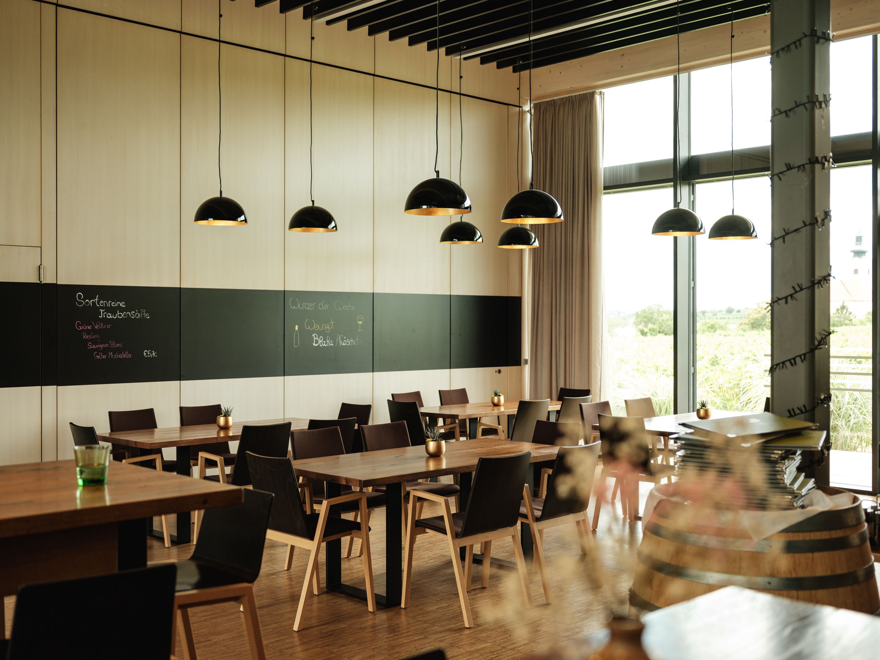 Modern restaurant with wooden tables, black chairs and large windows. Black hanging lamps and a blackboard on the wall.