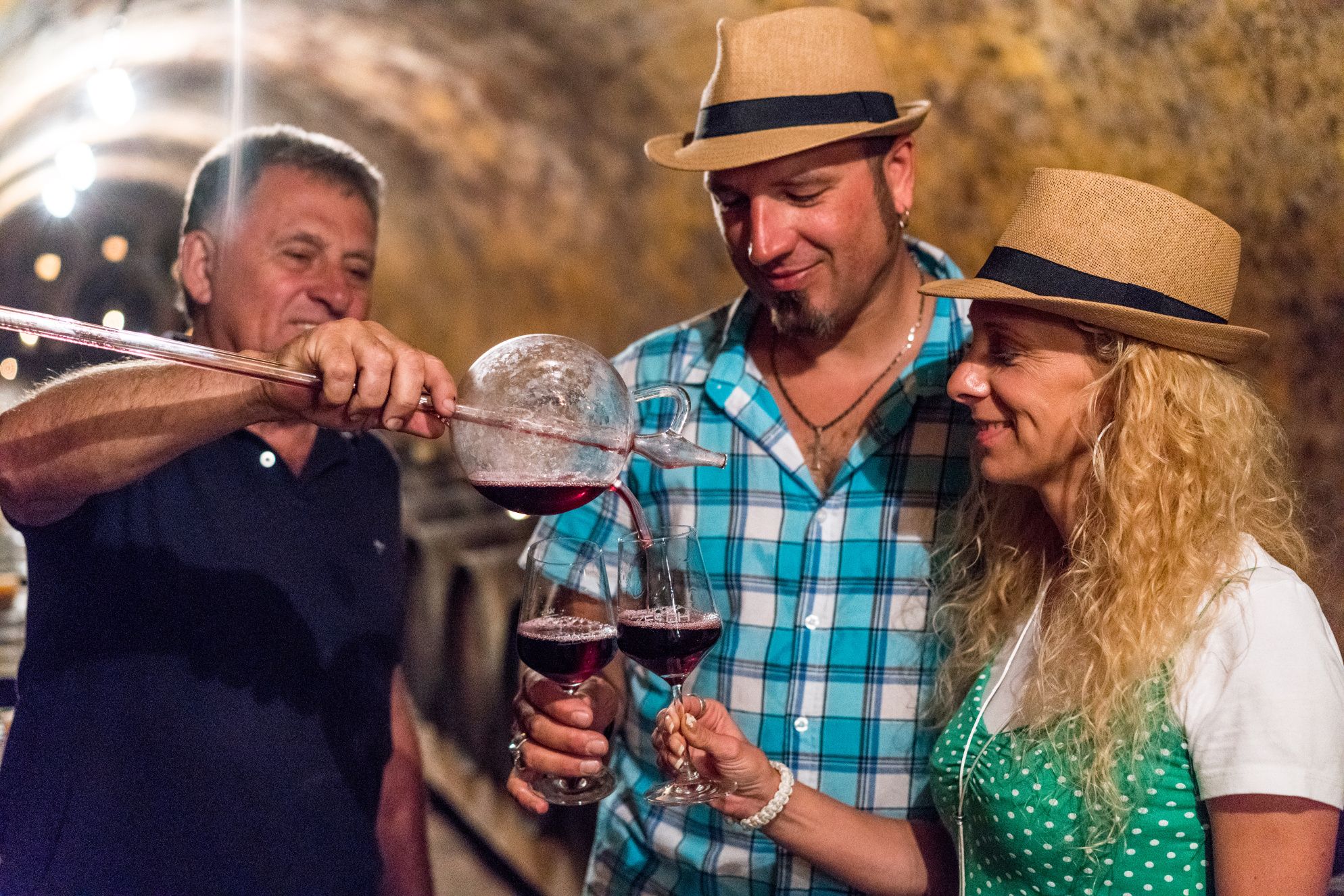 Three people in a wine cellar, two wearing straw hats. A man is pouring red wine into glasses.