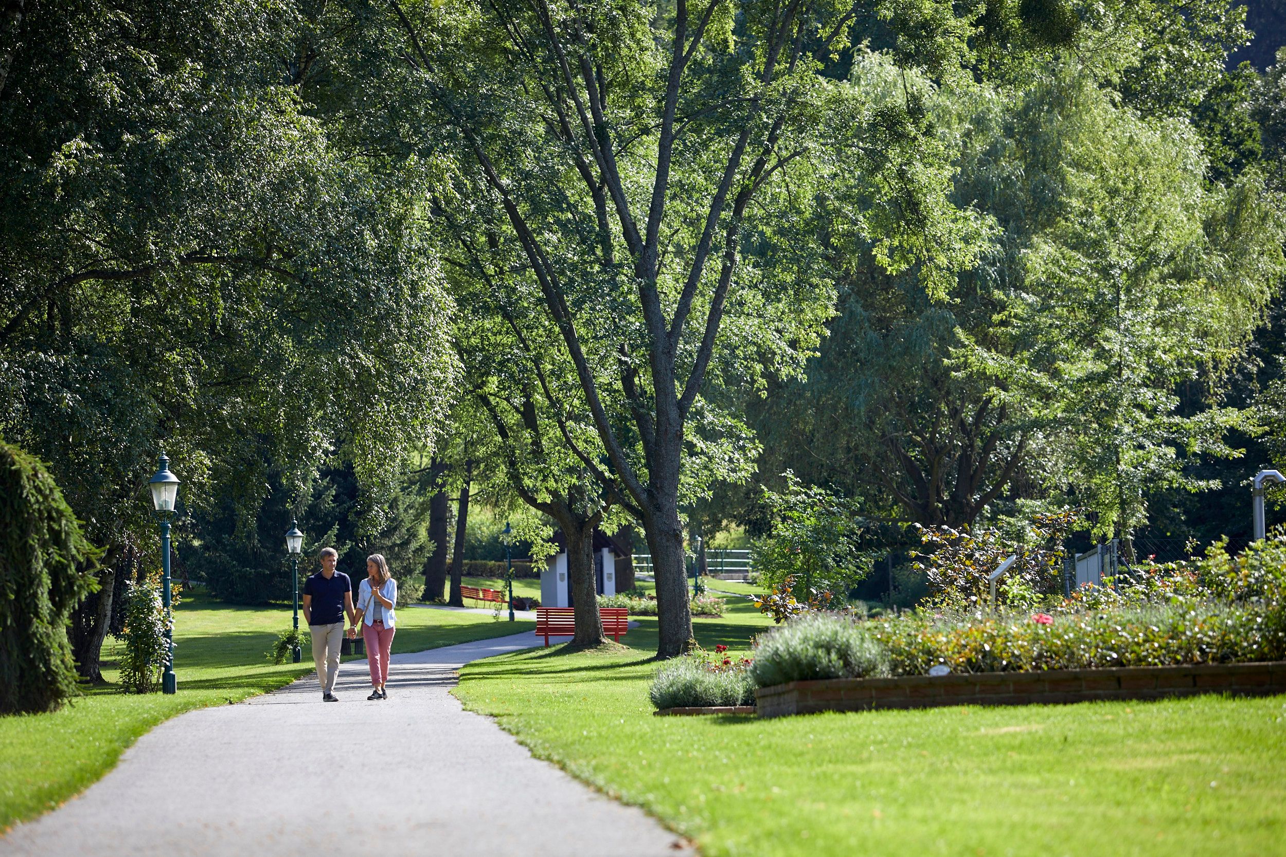 A couple walks along a path in the spa gardens of Bad Schönau, surrounded by green trees and well-tended flowerbeds.