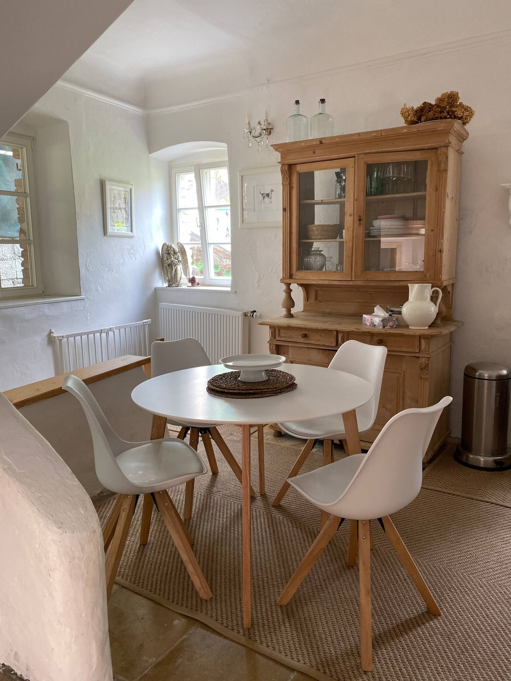 Dining area with round table, four white chairs, wooden cupboard and decoration in a bright room.