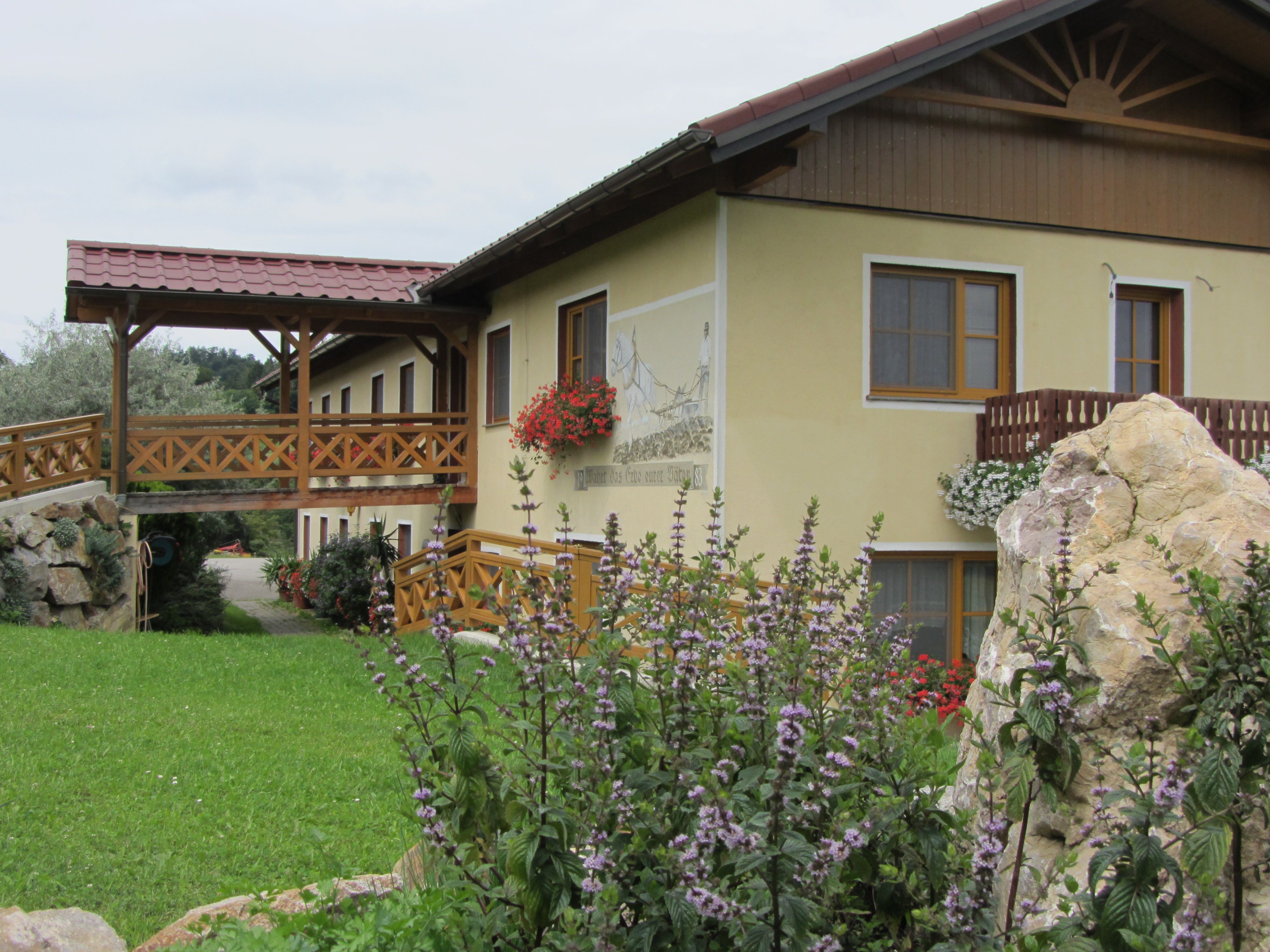 A yellow building with a wooden balcony and flowers, surrounded by green countryside.