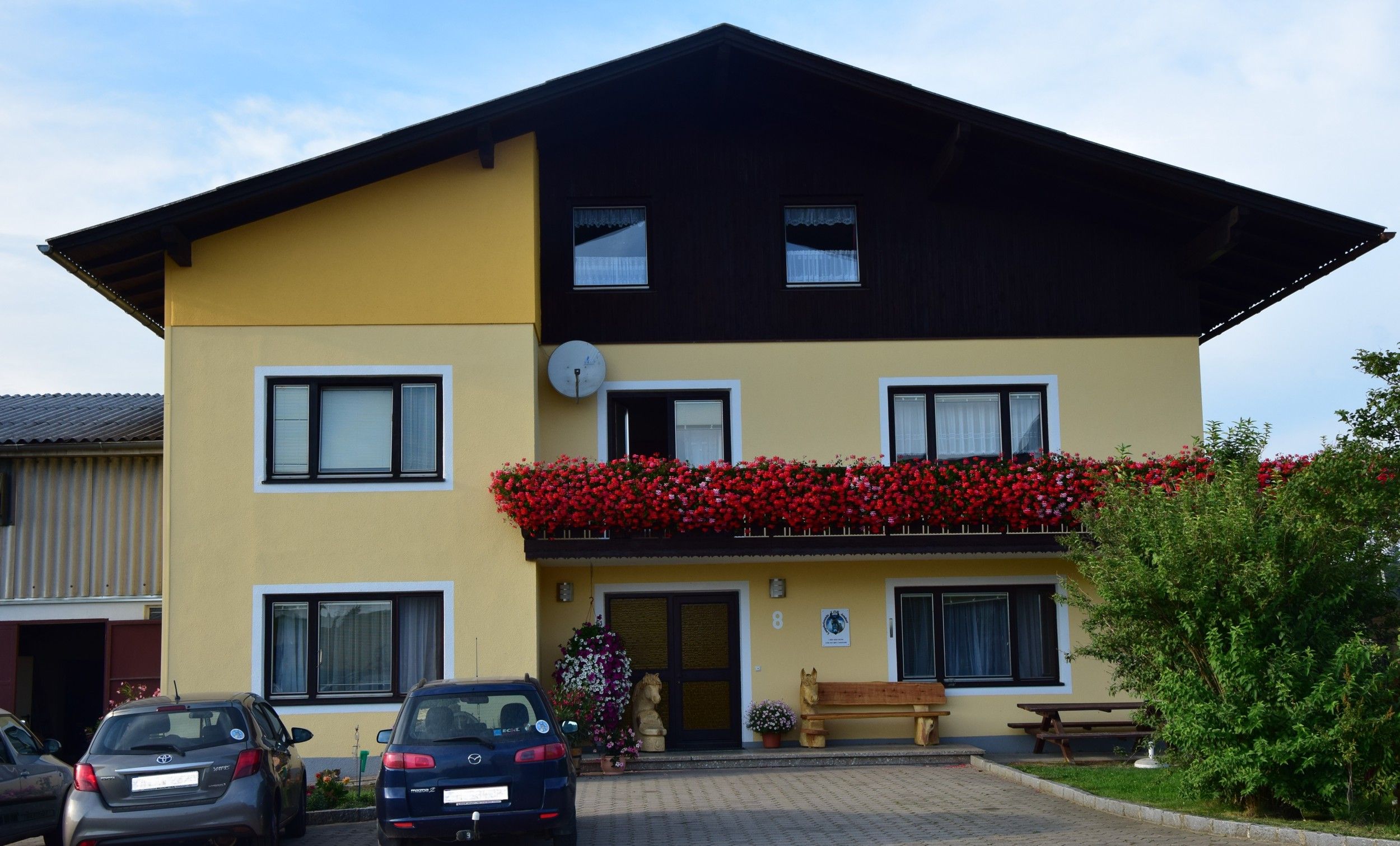 Exterior view of a yellow house with a balcony full of red flowers and parked cars in front of it.