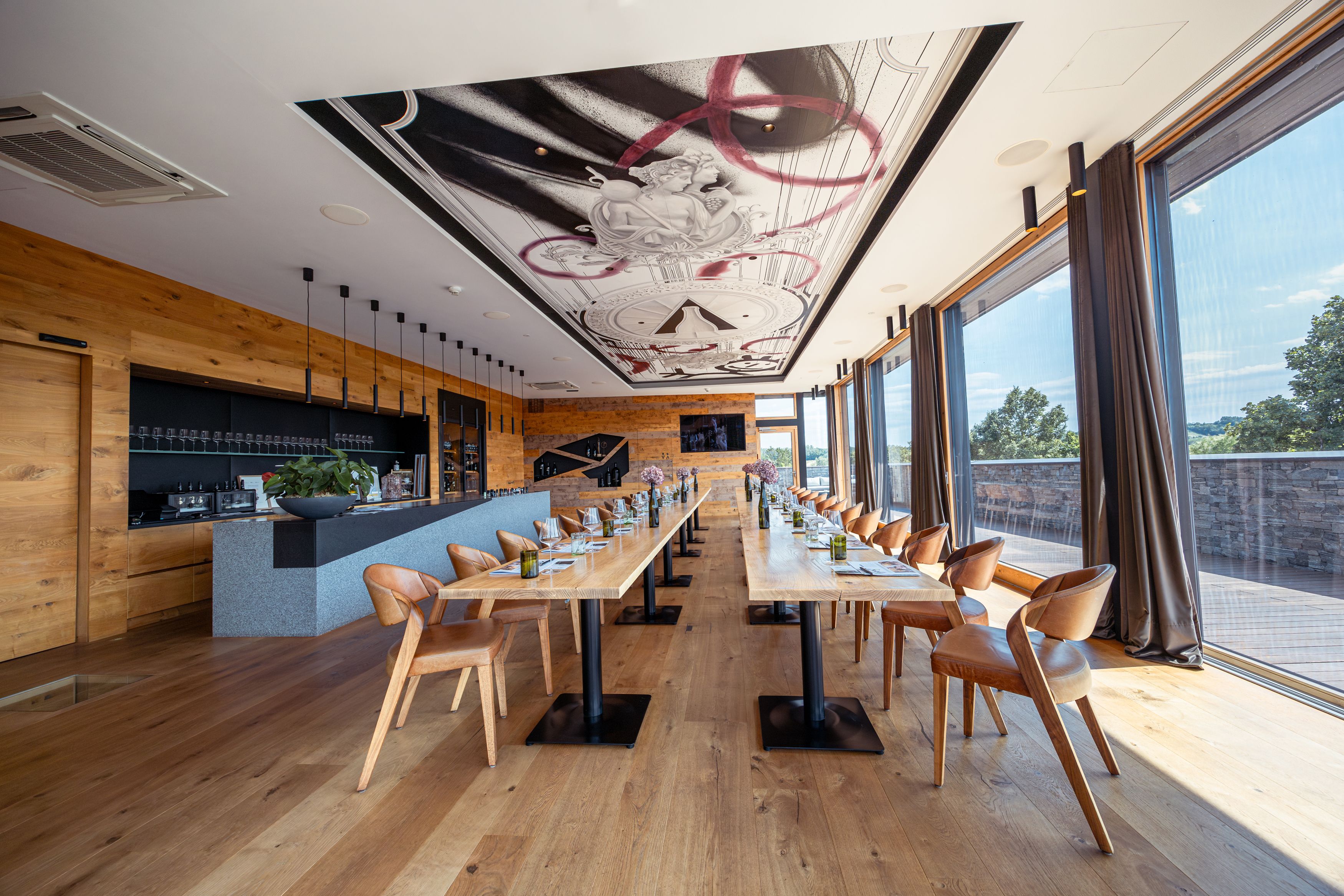 Interior view of a modern restaurant with long wooden tables, leather armchairs and large windows.