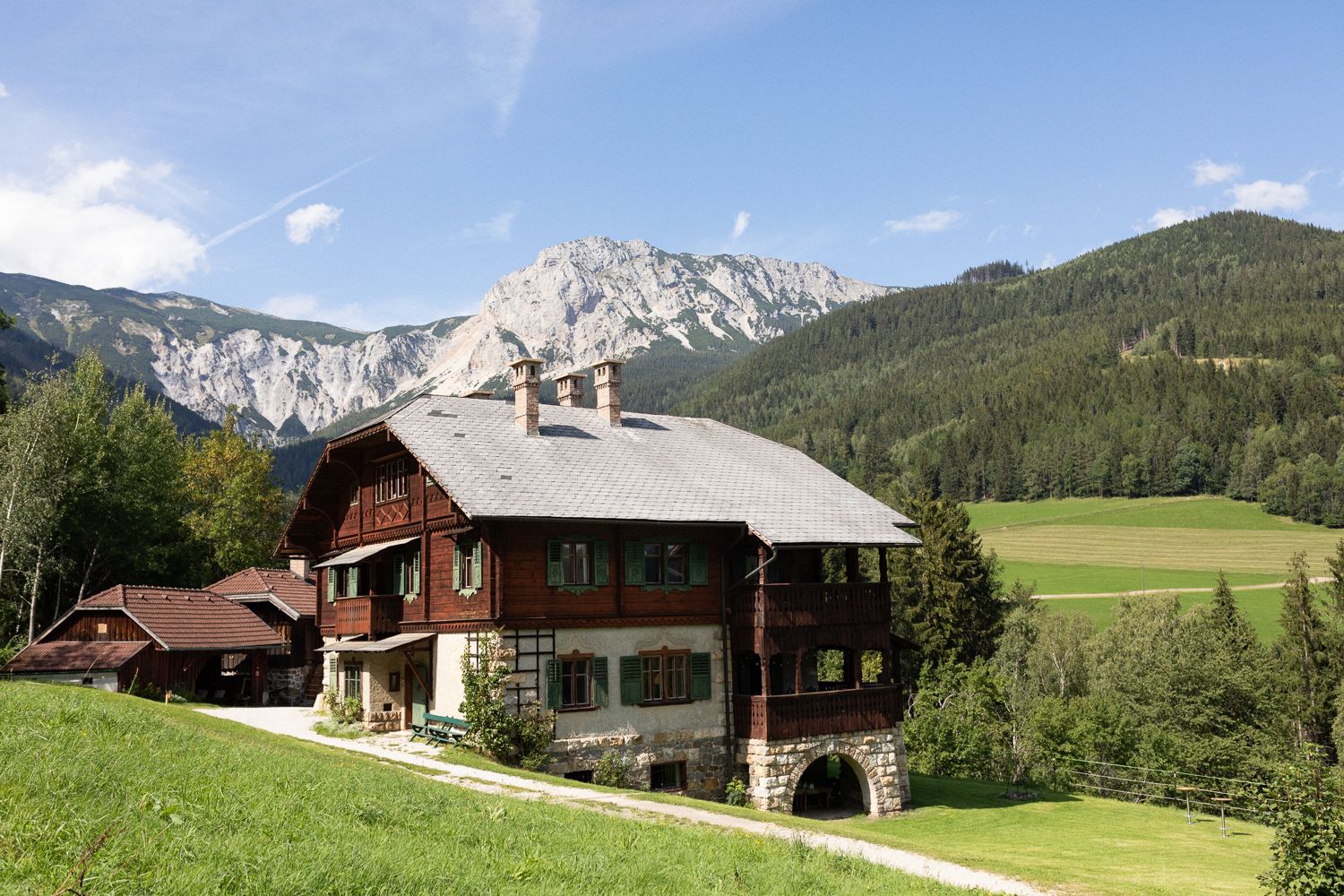 A traditional wooden house in a mountainous landscape with meadows and forests in the background.