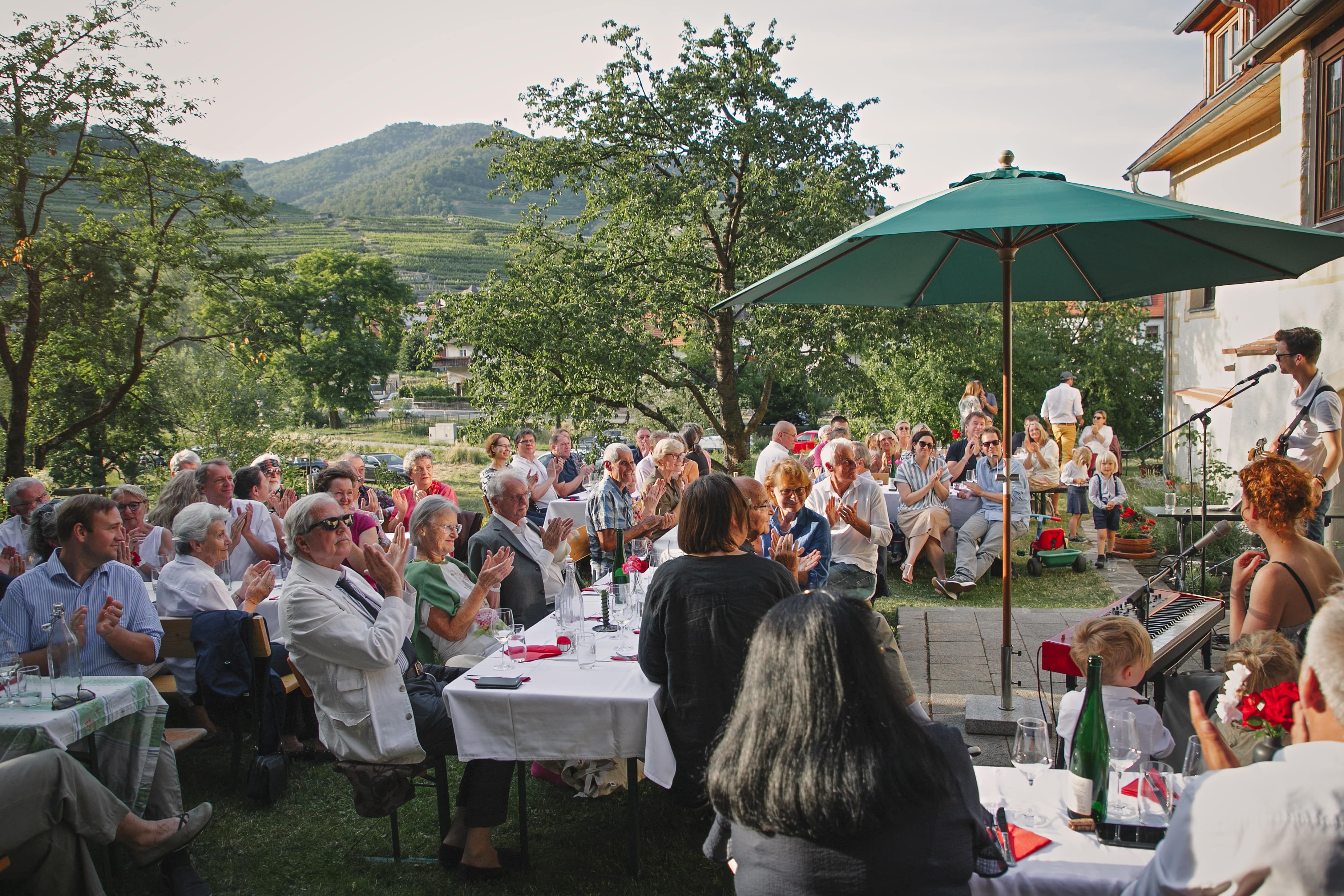 People sitting outside at an event with music, surrounded by trees and mountains.