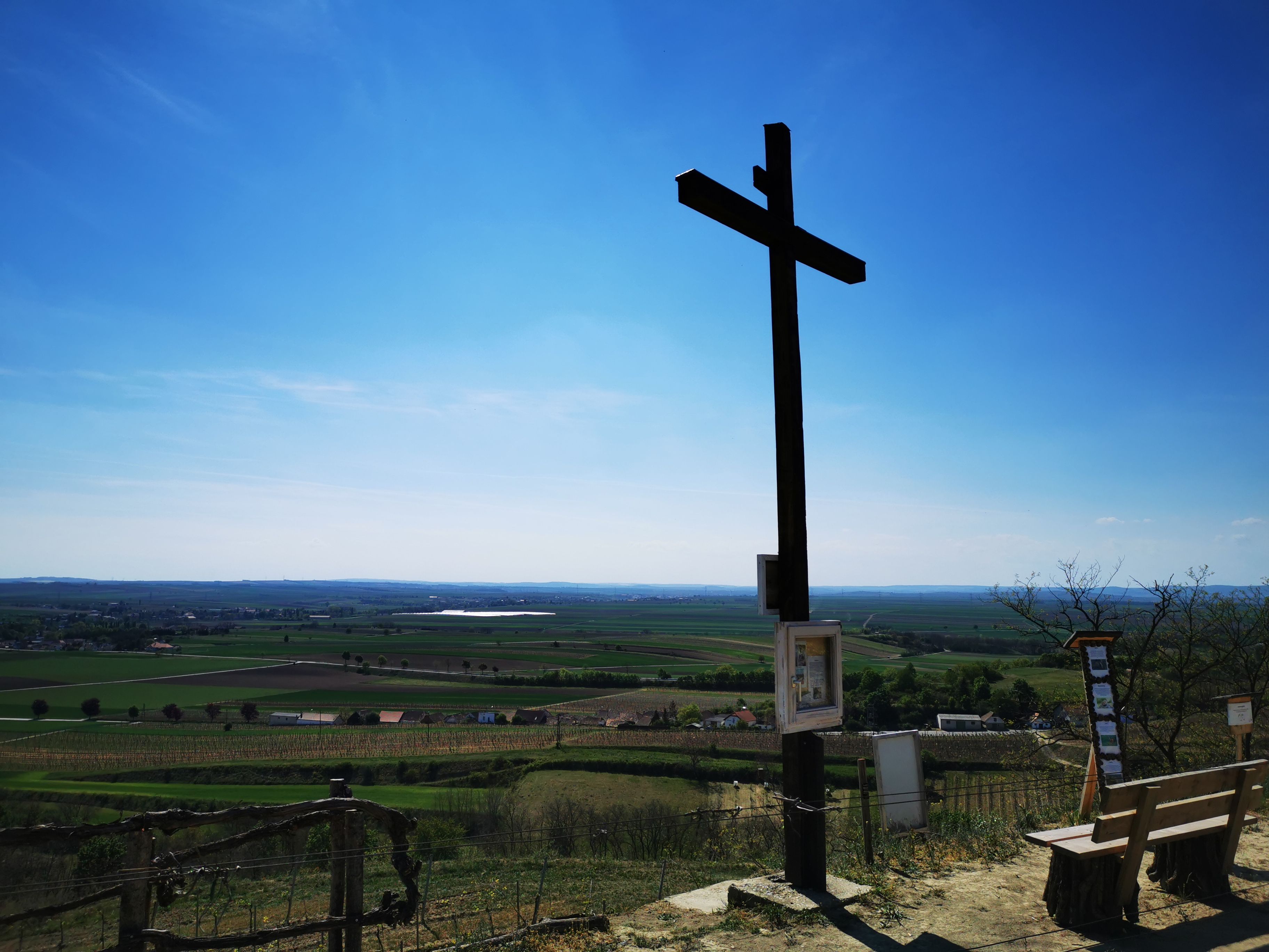 A large wooden cross on a hill overlooking a vast landscape under a blue sky.