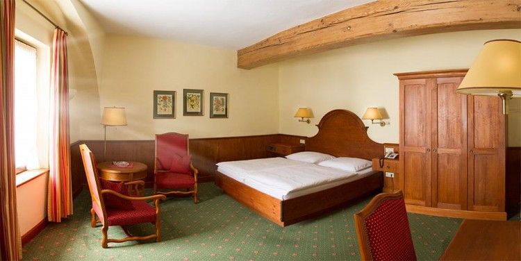 Cozy hotel room with wooden furniture, double bed, two red armchairs and a wooden table. Wooden beams on the ceiling, green carpets and murals.
