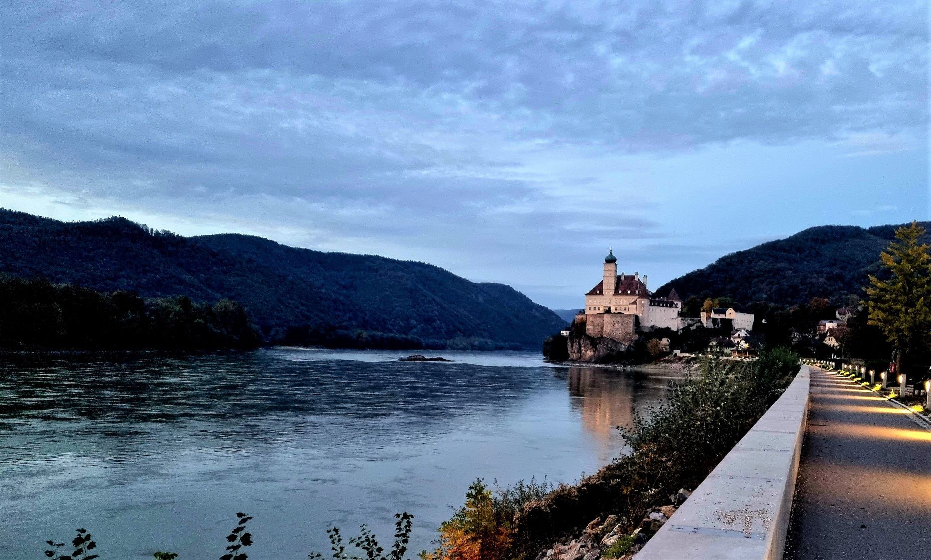 An illuminated path leads along a river to a castle on a hill surrounded by wooded mountains.