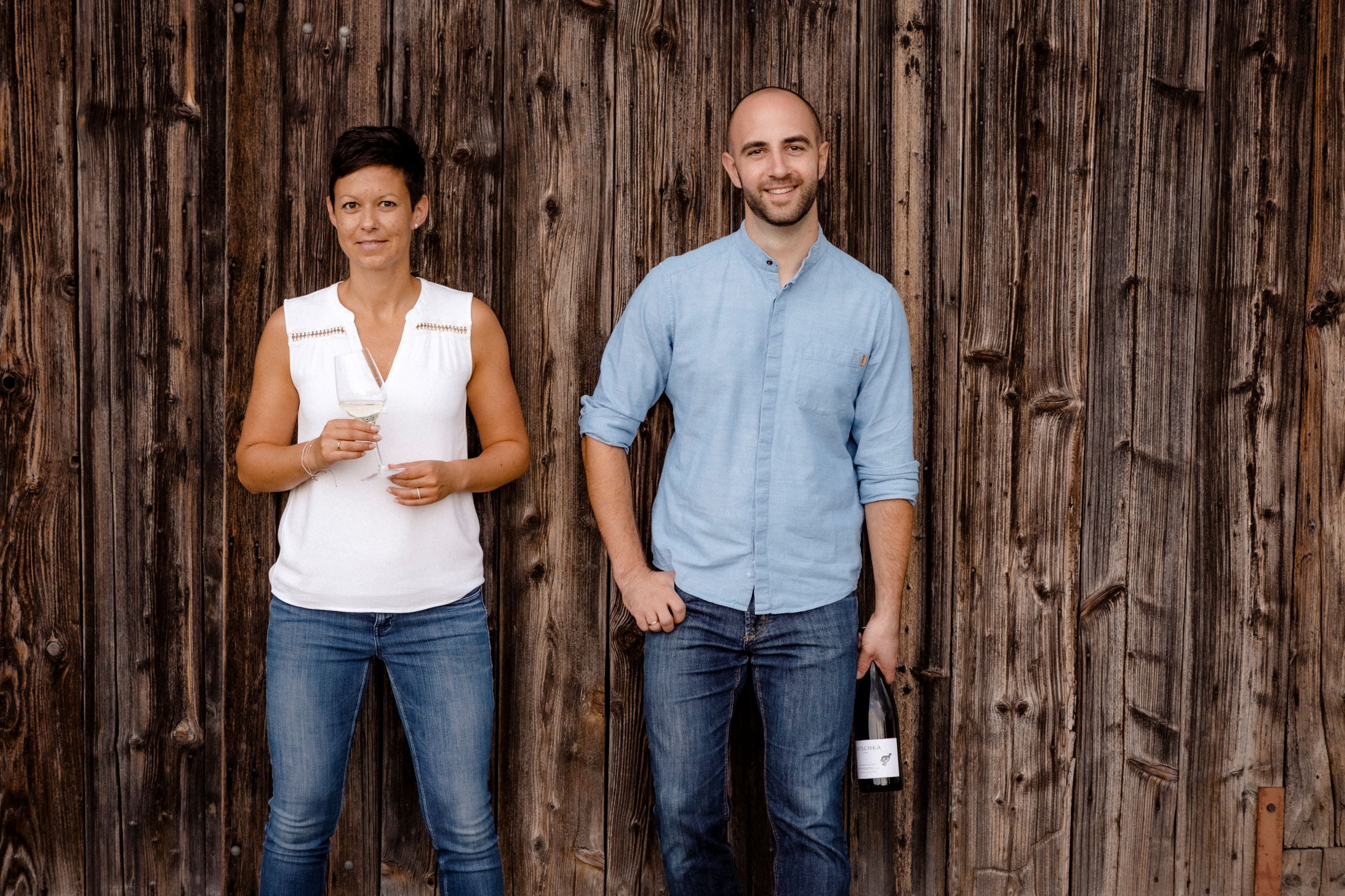Two people are standing in front of a wooden wall, a woman with a wine glass and a man with a wine bottle.