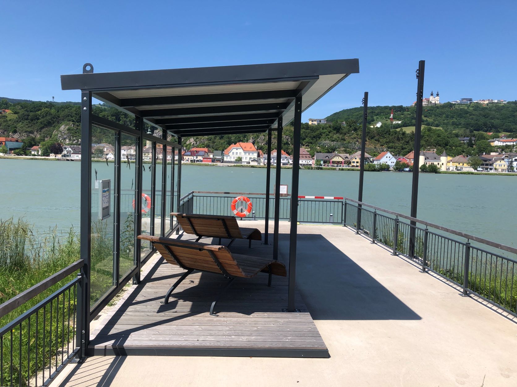 Landing stage with benches by the river, a town and a basilica on a hill in the background.