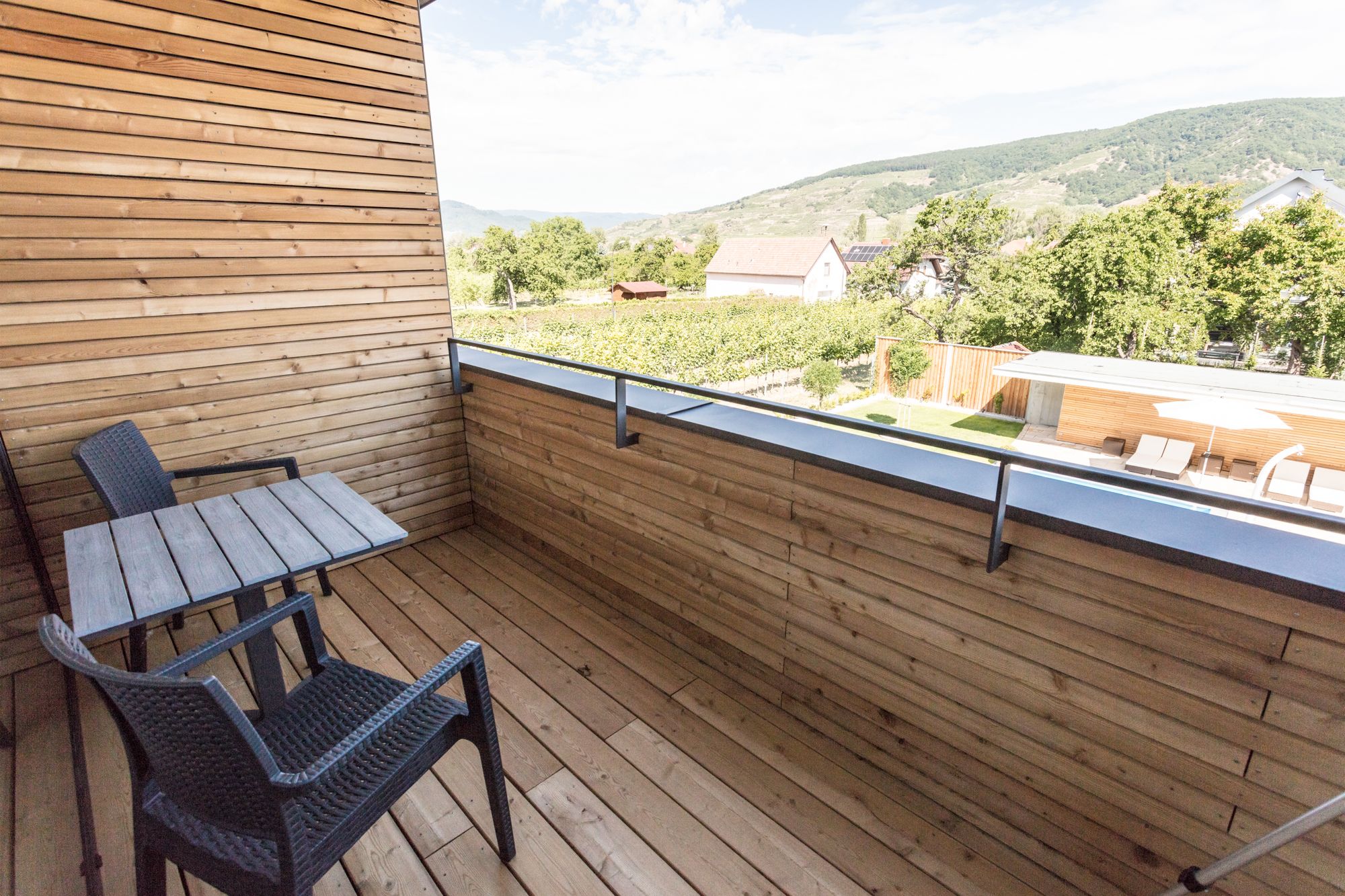 Wood-paneled balcony with table and two chairs, view of green landscape and hills in the background.