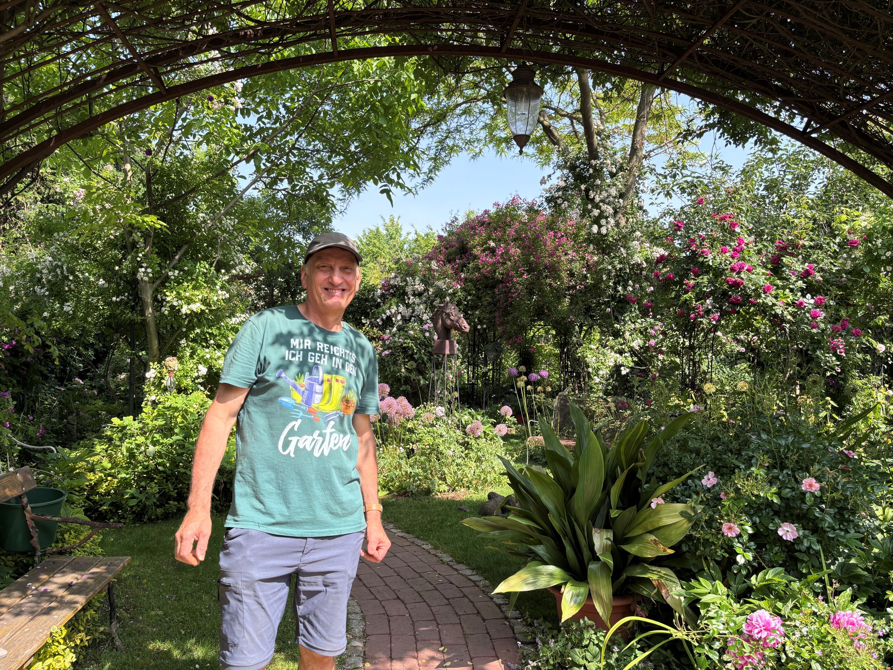A man stands in a flowering garden under a pergola.