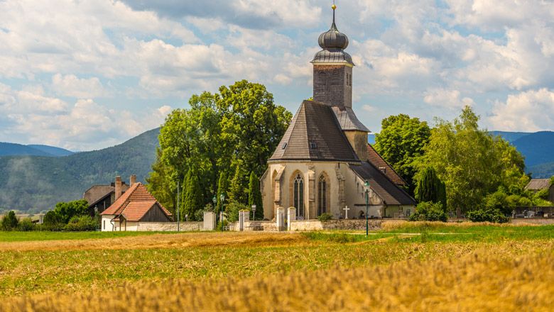 Church in Muthmannsdorf surrounded by fields and trees, with mountains in the background.