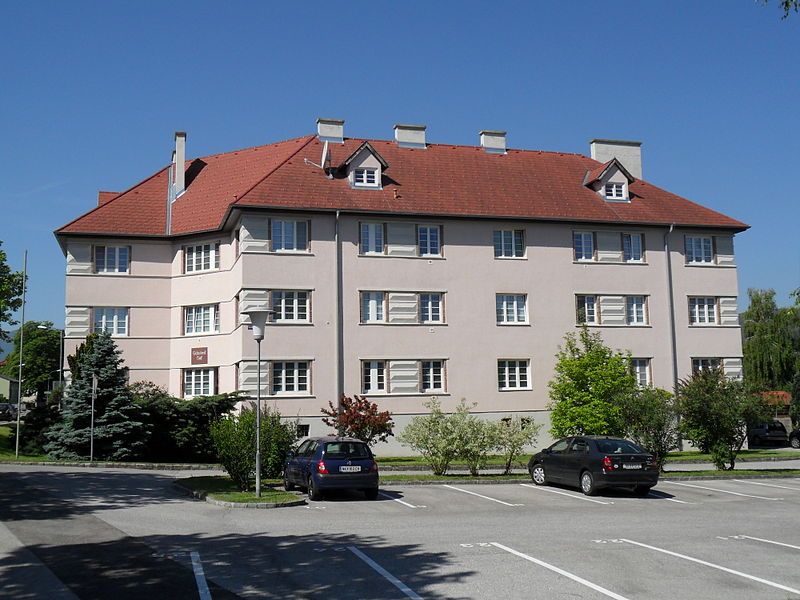 Residential building with a red roof and several windows, surrounded by trees and parked cars.