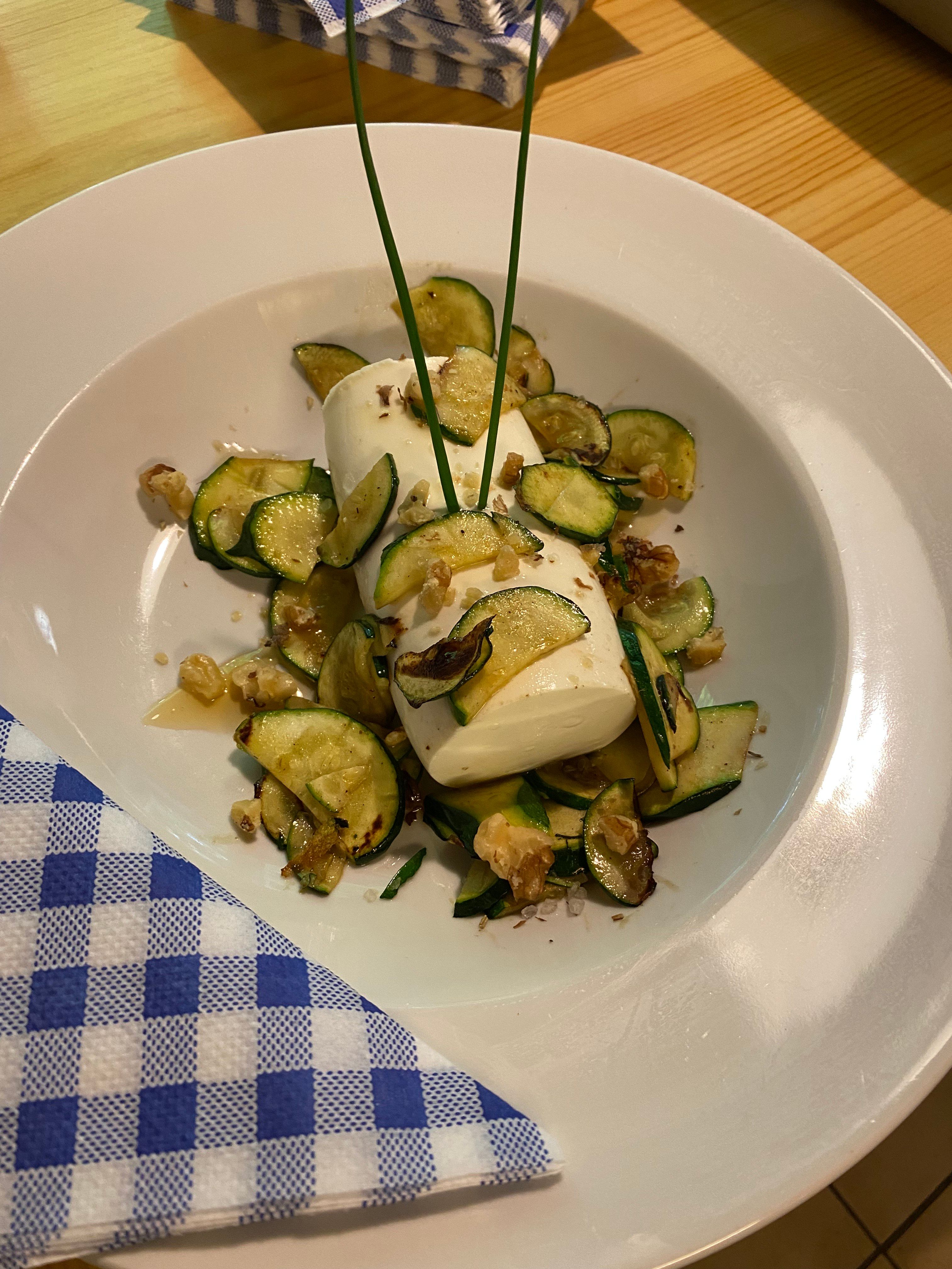 A plate with zucchini slices, mozzarella and walnuts, decorated with chives, on a wooden table with blue and white checkered napkins.