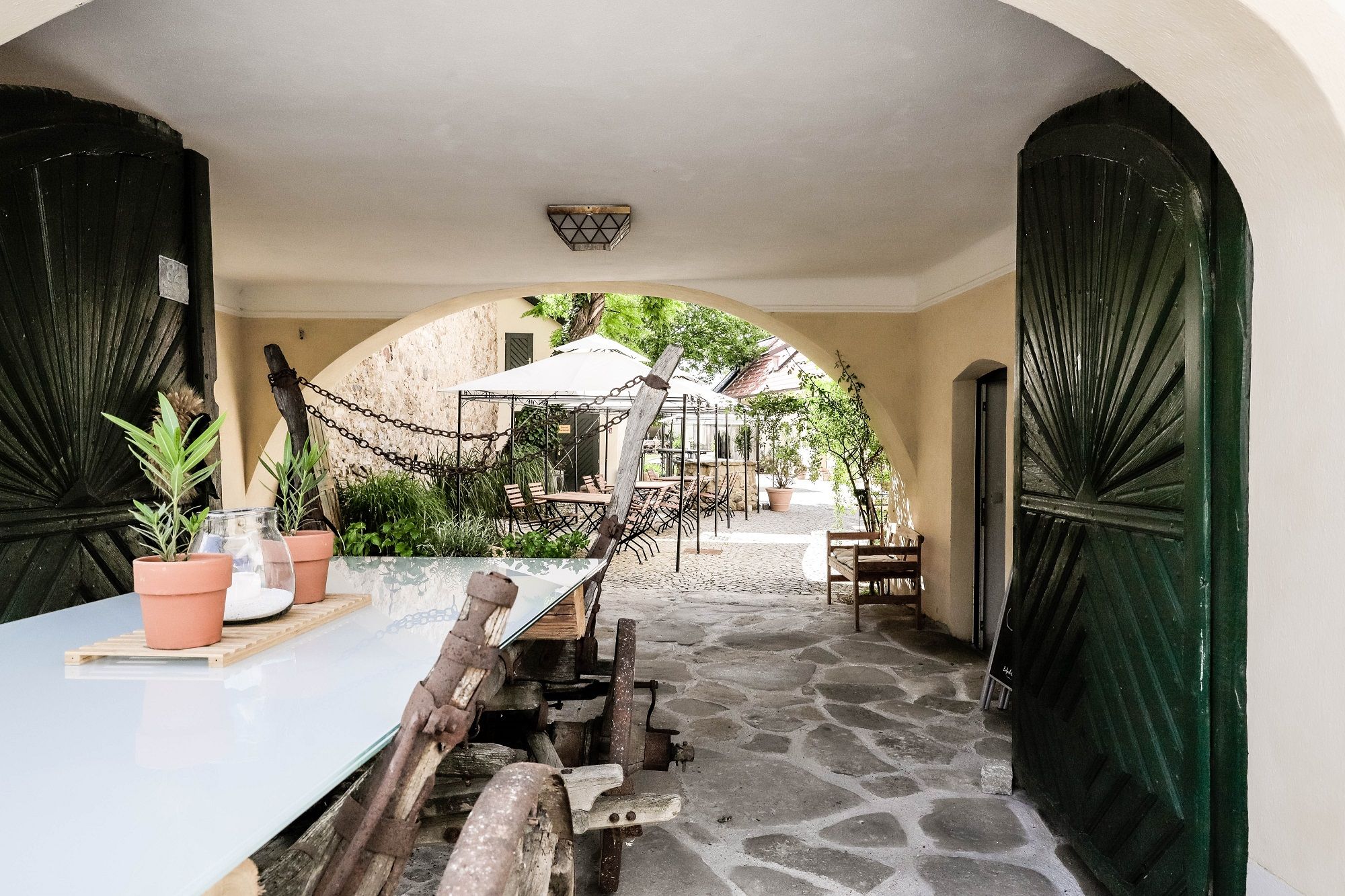 A covered passageway with a wooden table and plants leads to a paved courtyard with tables and chairs.
