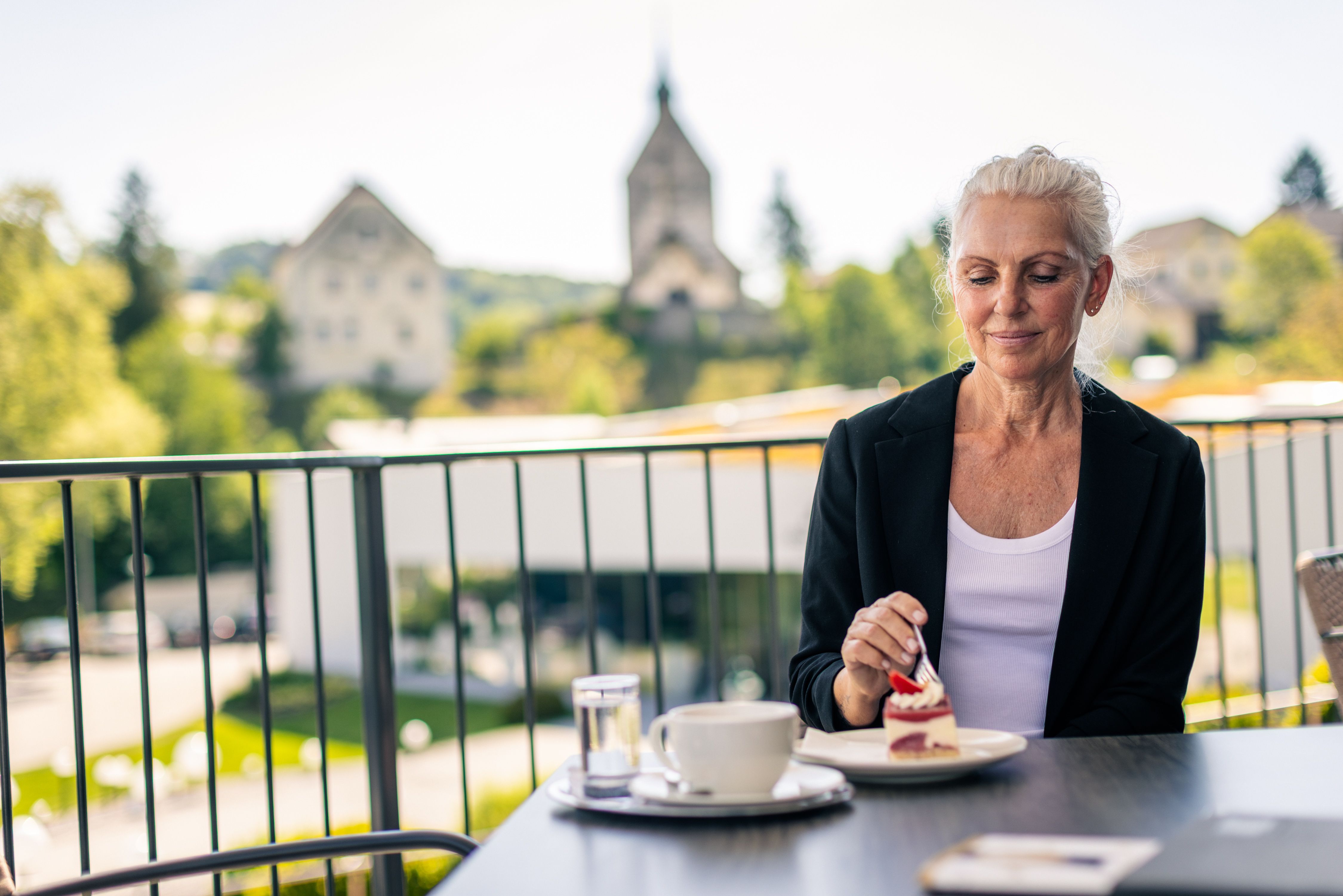 A woman is sitting on a terrace, enjoying a slice of cake with a cup of coffee. The fortified church in Bad Schönau can be seen in the background.