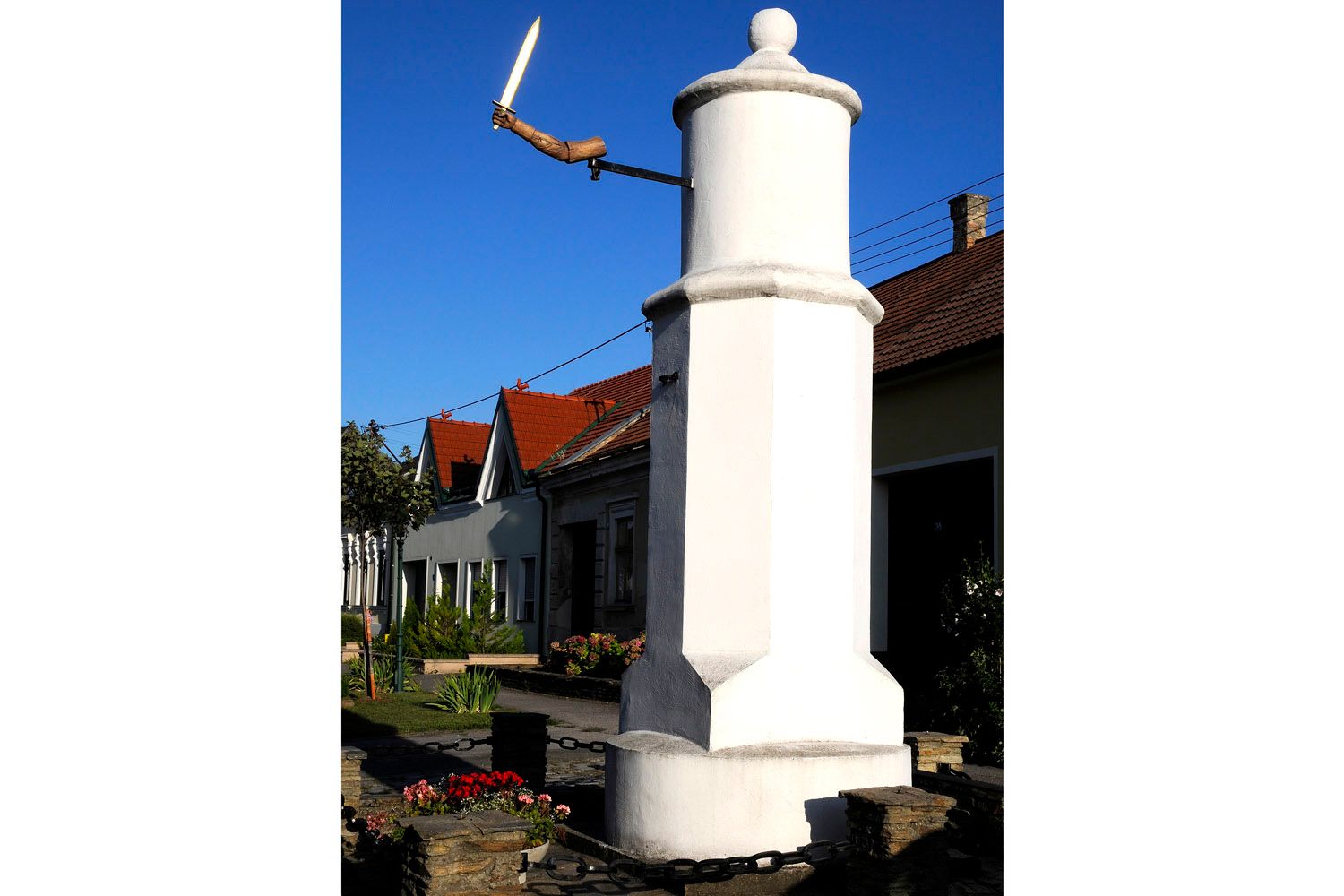A white pillory with a wooden hand holding a sword in front of a house with red roofs.