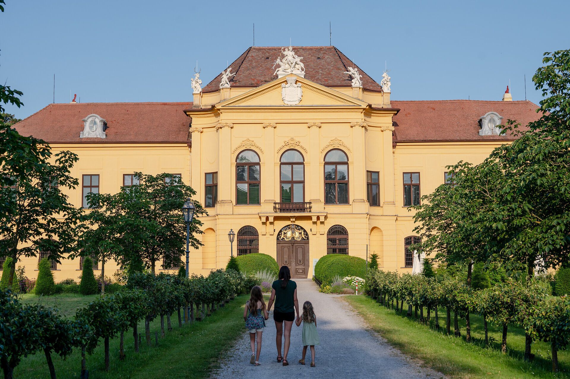 Woman holding the hands of two girls walking along a gravel path towards Eckartsau Castle. 