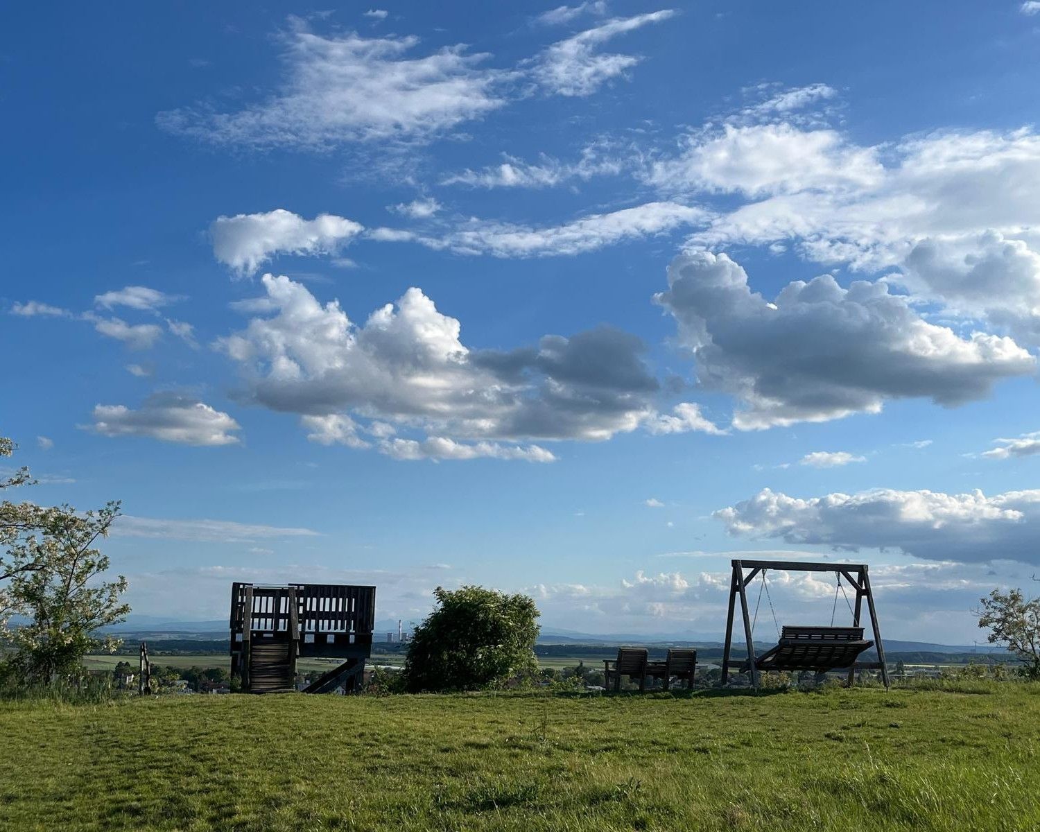 A wide sky with large white clouds over a green meadow. In the foreground are a wooden platform, several seats and a large wooden swing with a view across the wide landscape to the horizon. The evening sun creates a calm, open atmosphere.