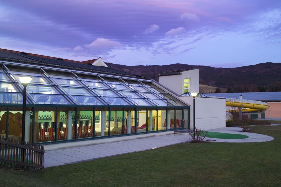 Exterior view of the Yspertal indoor pool at dusk with glass façade and slide.