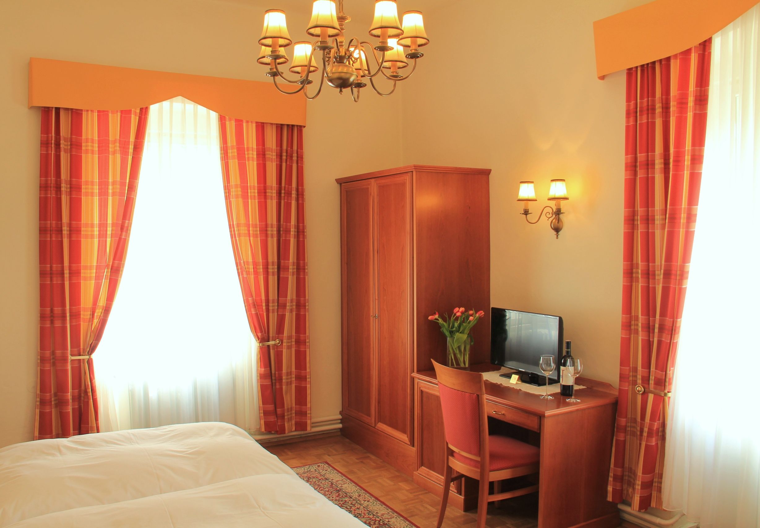 A cozy hotel room with a double bed, wooden furniture, a desk with TV and a vase of flowers. Curtains in red-orange.