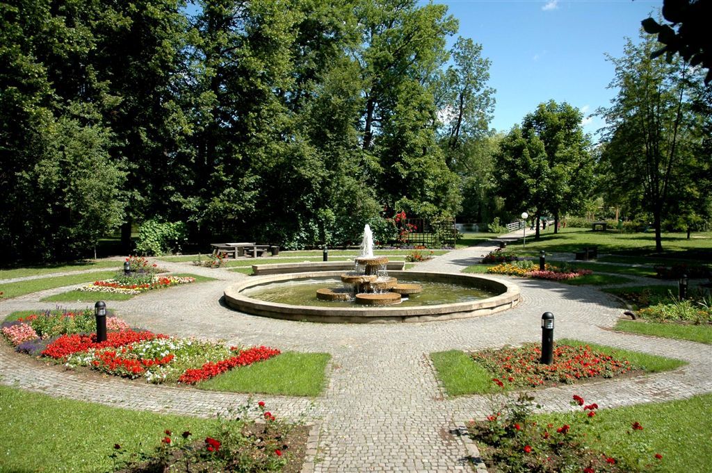 A well-kept park with a central fountain, surrounded by flowerbeds and trees.