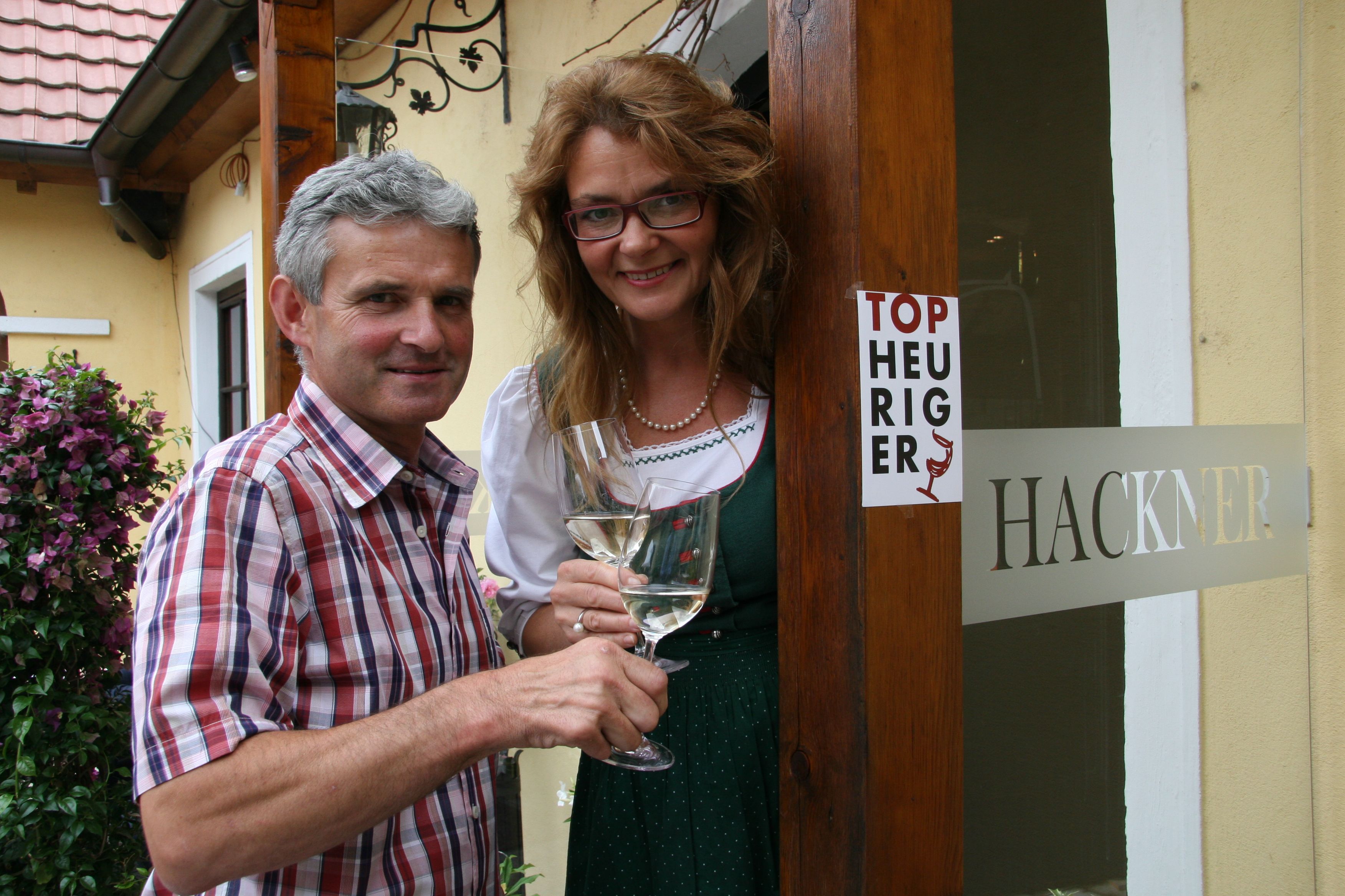 A man and a woman are standing in front of a building with a sign saying 'Top Heuriger' and holding wine glasses.