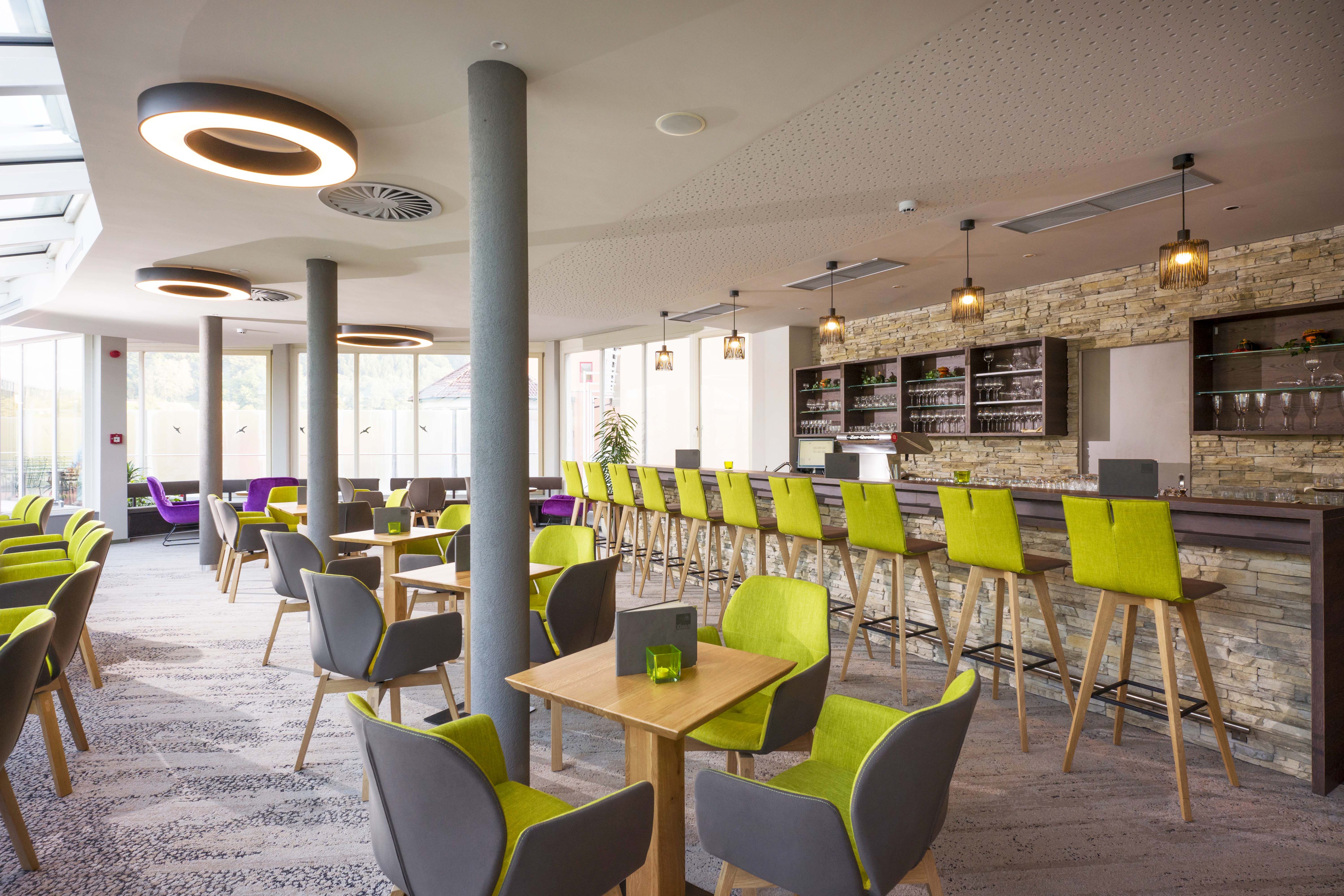 Modern café with green and gray chairs, wooden tables and a long bar with a stone wall.