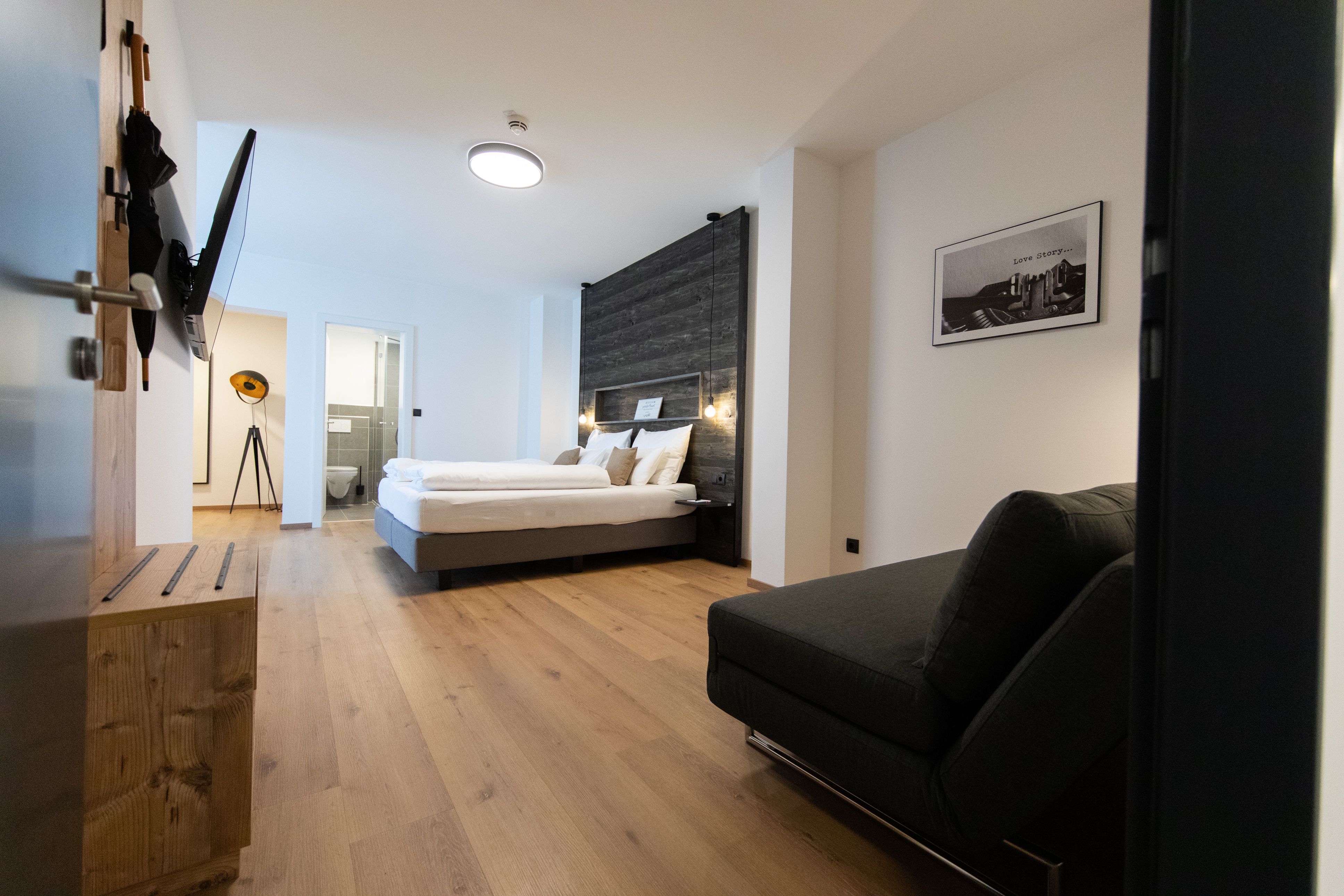 Modernly furnished hotel room with double bed, sofa and flat-screen TV. Light wooden floors and a bathroom in the back.