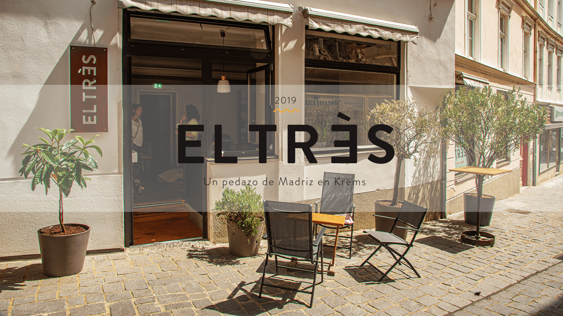 Exterior view of a café called EL TRÈS, seating and plants in front of the entrance.