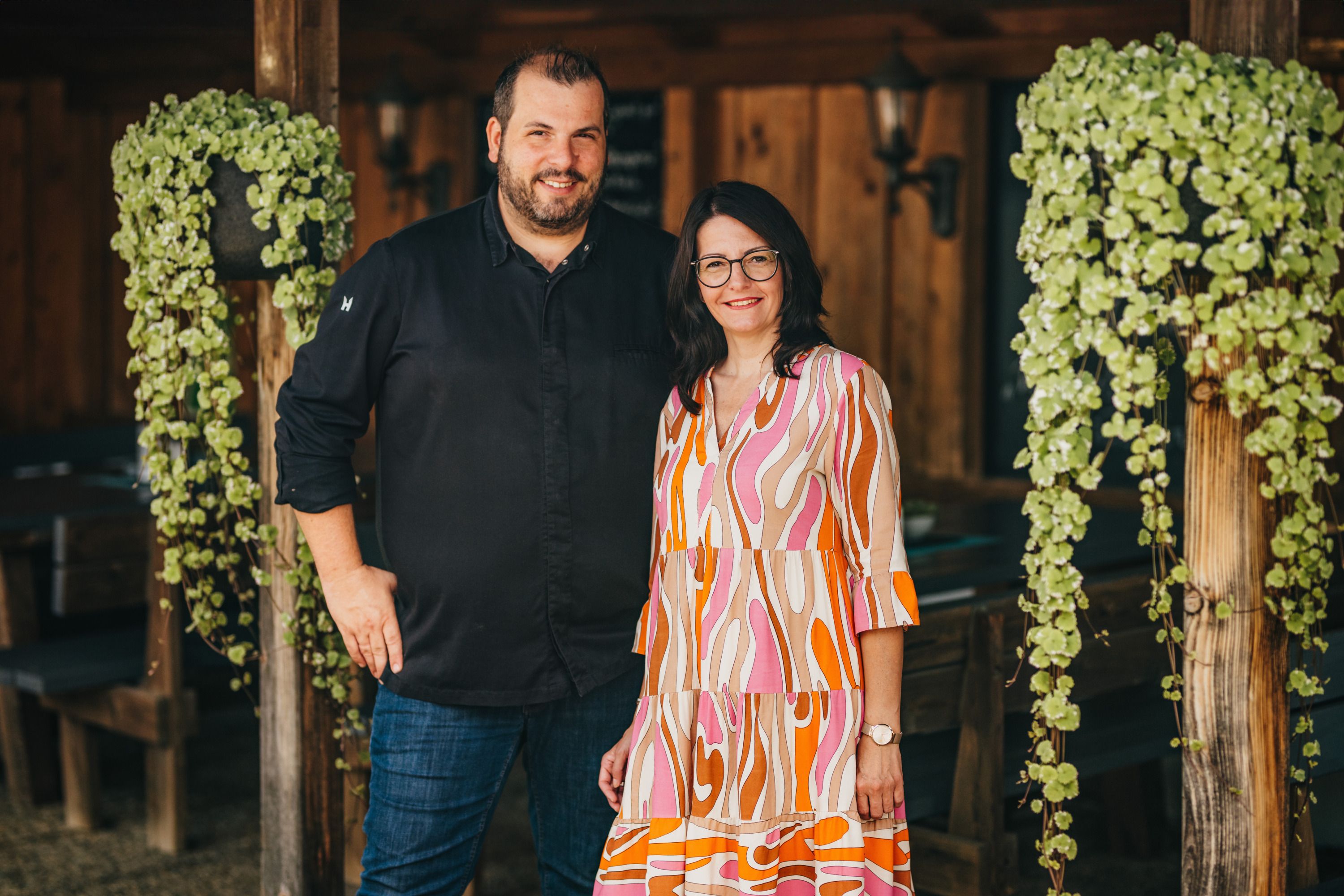 A man and a woman stand smiling next to each other in a rustic room with wooden walls and hanging plants.