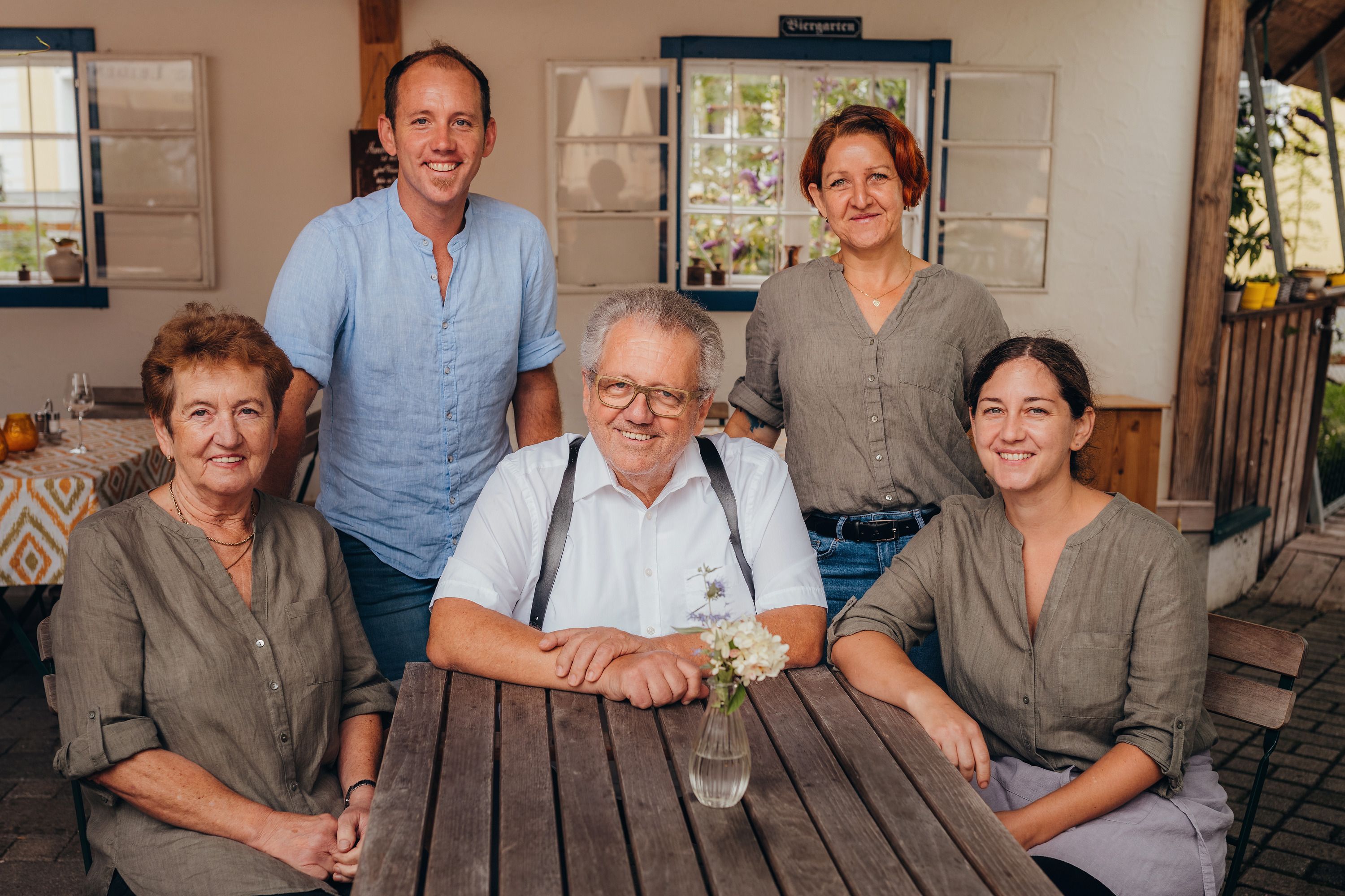 Five people sit and stand around a wooden table in a covered outdoor area.