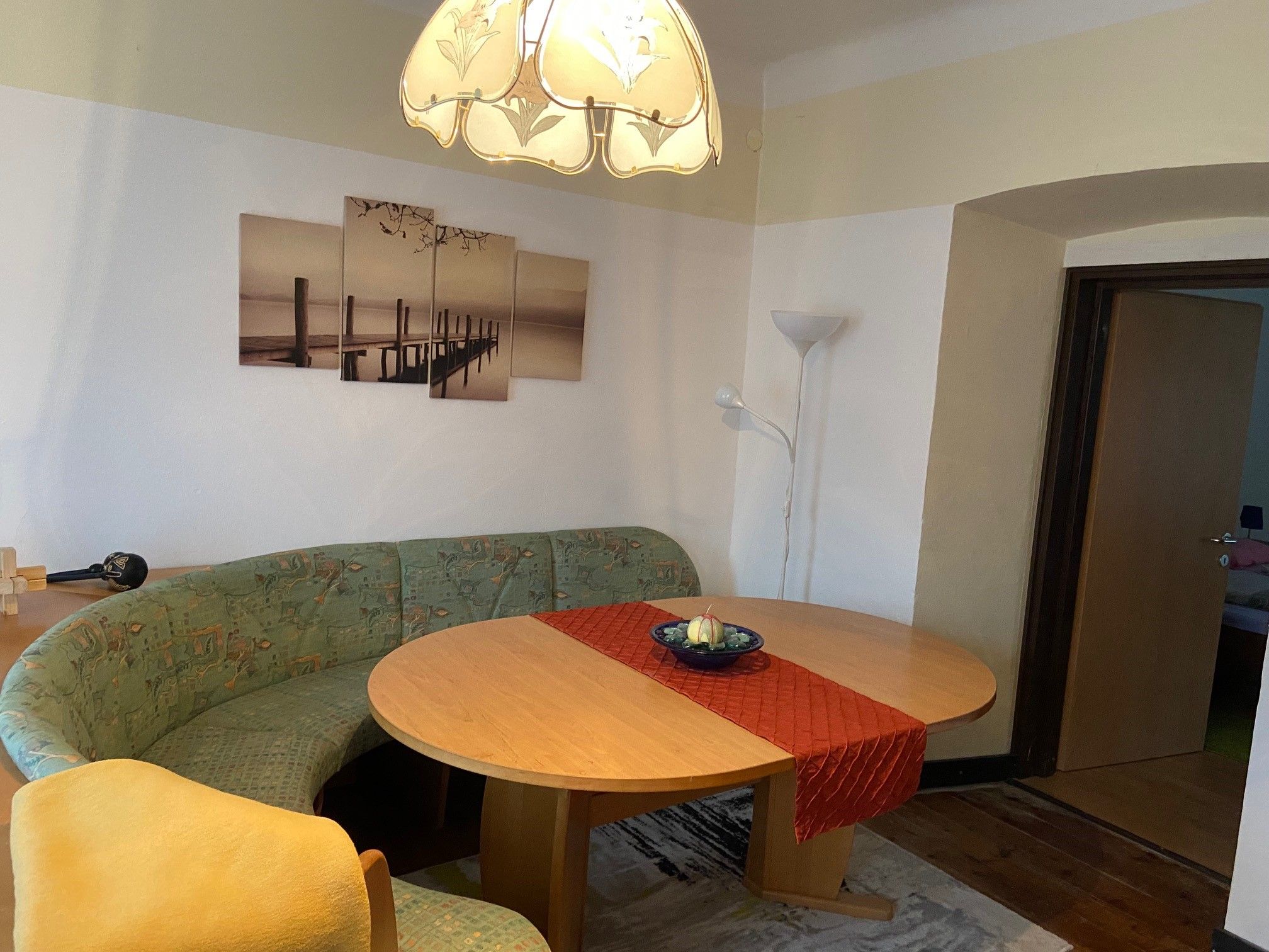 A living room with an oval wooden table, a green corner bench, a red table runner and a picture on the wall. A floor lamp stands in the corner.