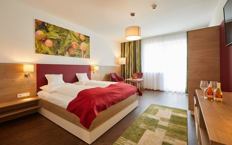 Modern hotel room with double bed, red ceiling, wooden furniture and large window. A picture of fruit hangs on the wall.