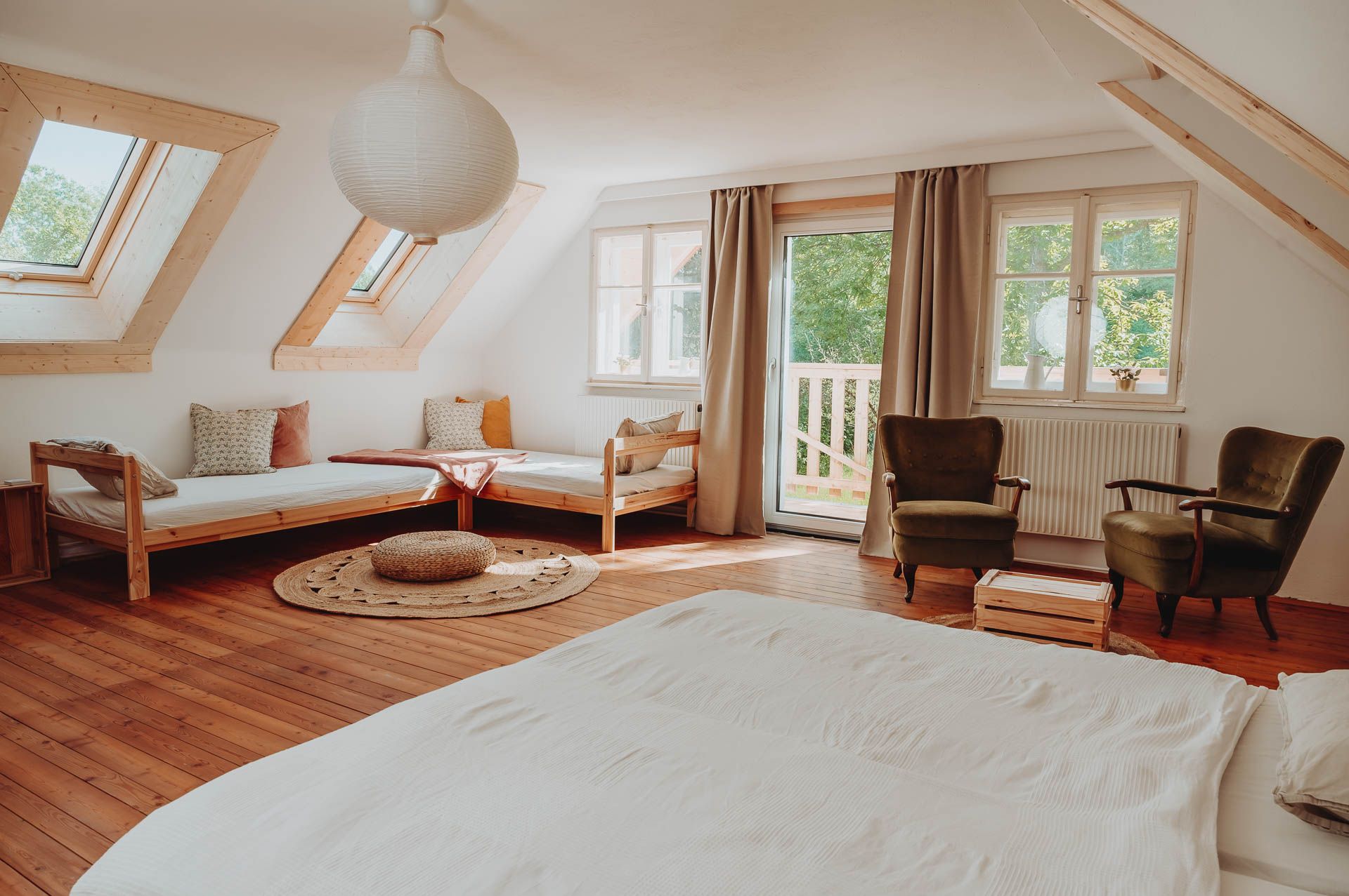 Bright multi-bed room with wooden floor, two beds, two armchairs and large windows.