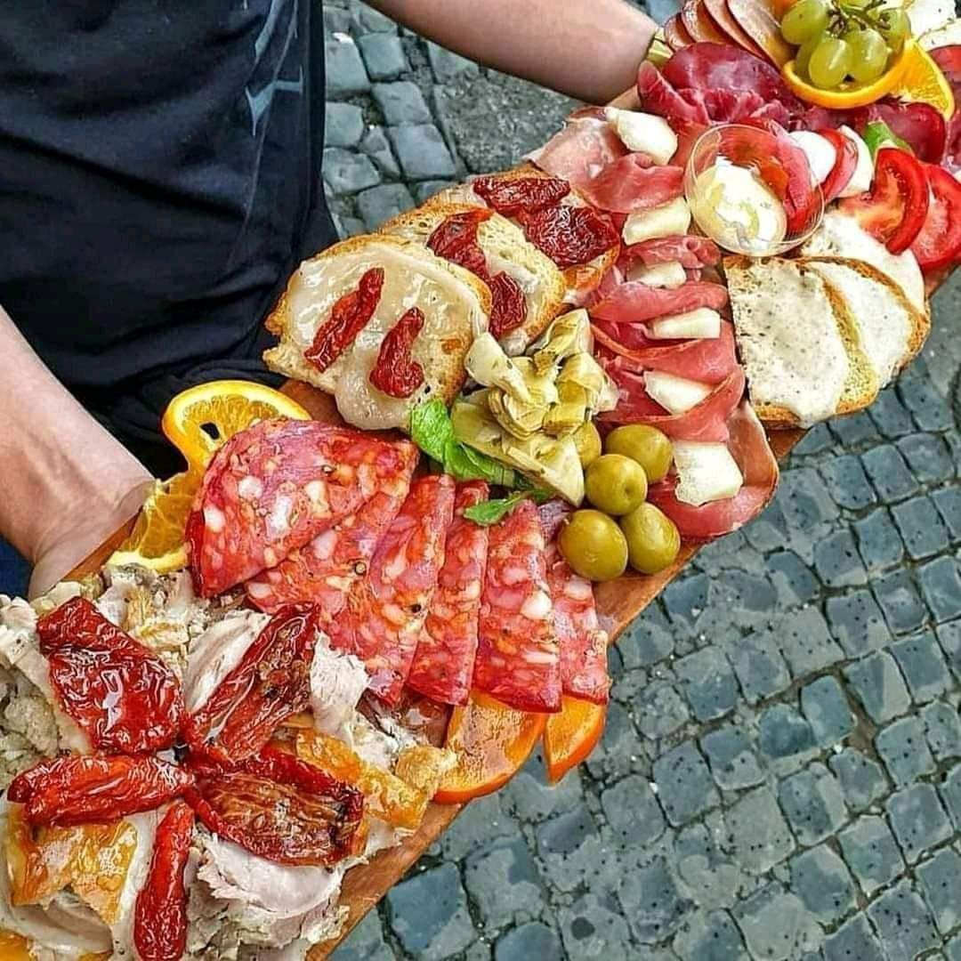 A large wooden platter with various antipasti, including sausage, cheese, olives and dried tomatoes, is held by one person.