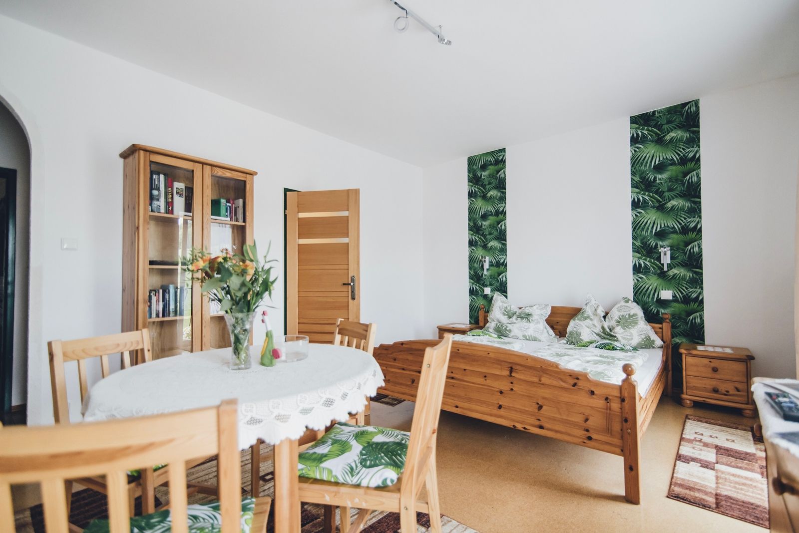 A cozy double room with wooden furniture, a table with flowers, a bed with a plant pattern and a bookshelf.