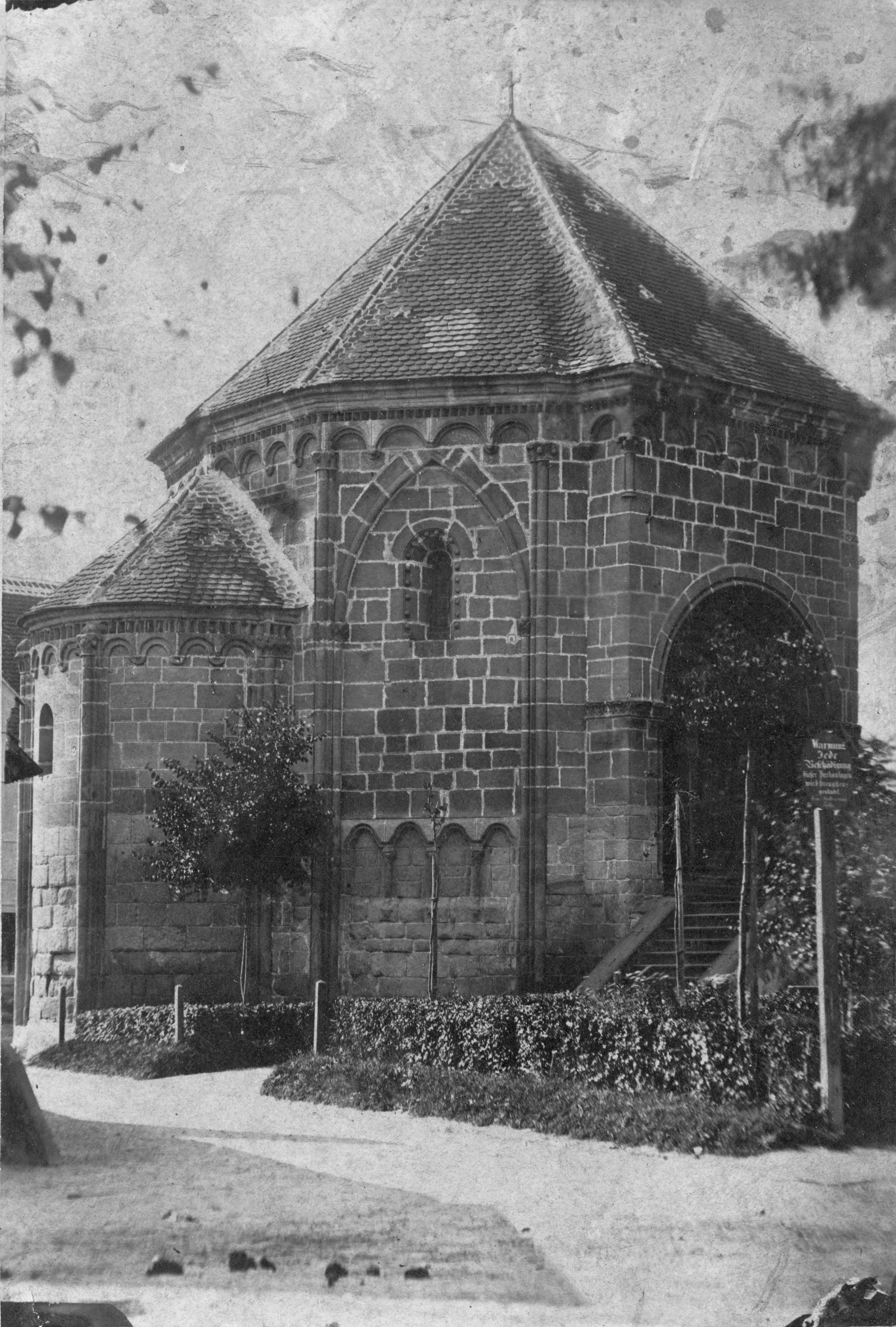 Historical photo of a late Romanesque charnel house with arched windows and tiled roof, surrounded by trees and a path.