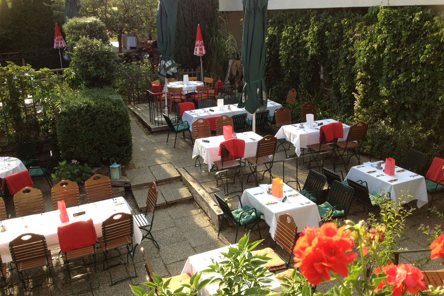 A cozy guest garden with covered tables, surrounded by green plants and red flowers.