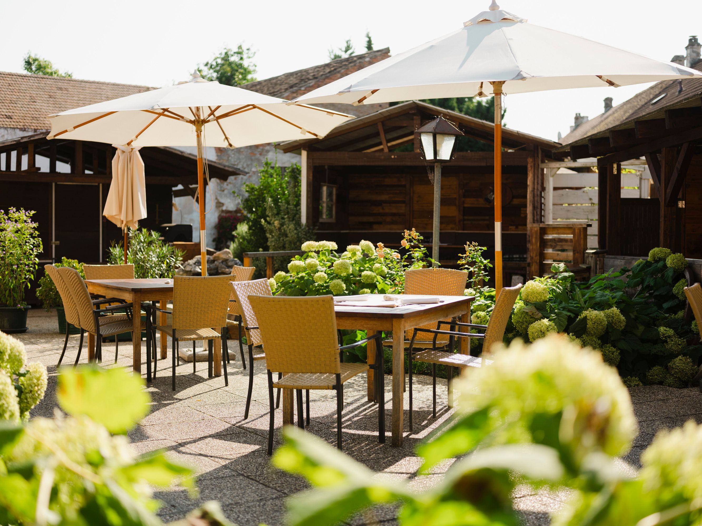 An idyllic guest garden with wooden tables, chairs and parasols, surrounded by green plants and flowers.
