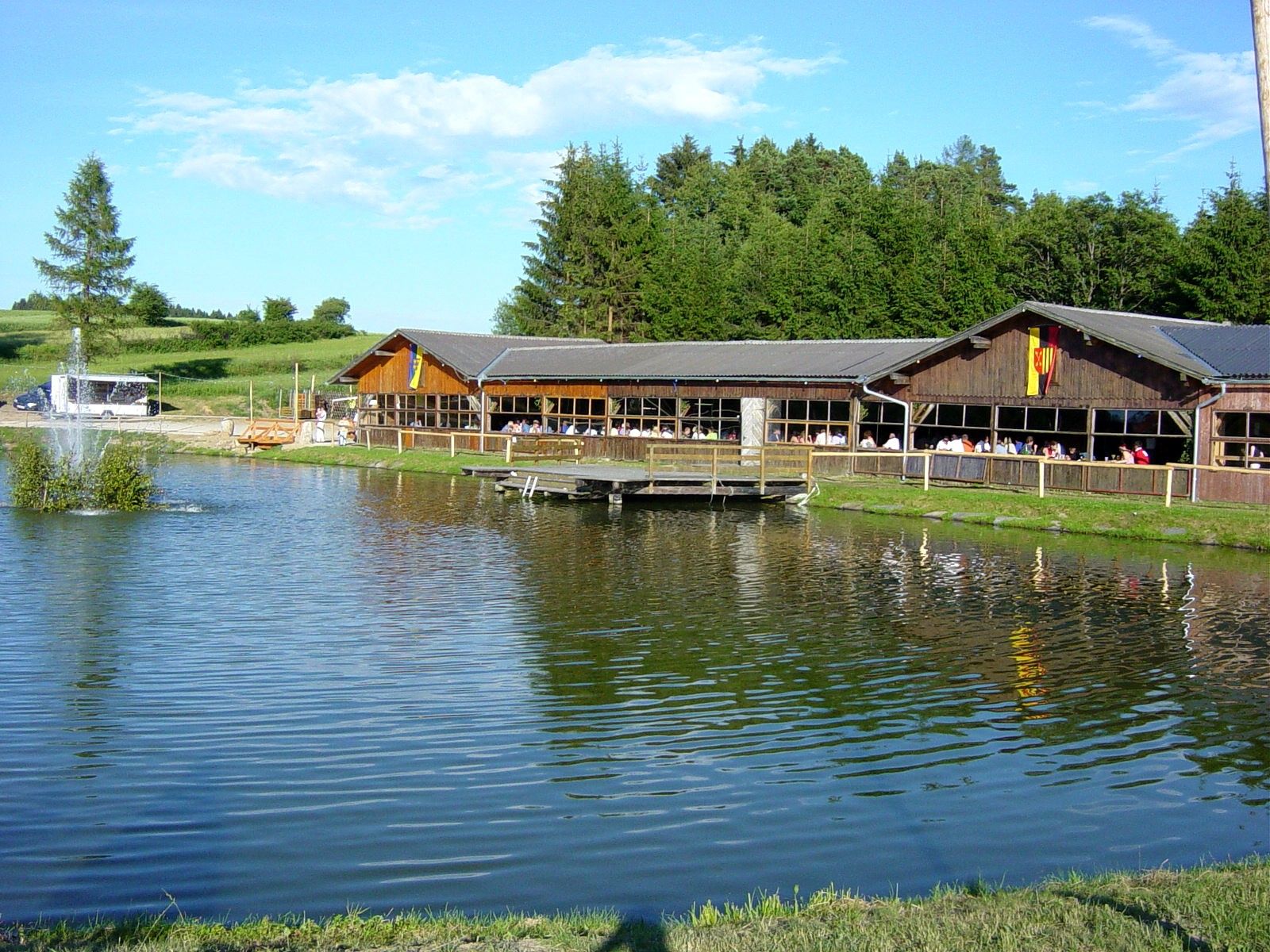 A wooden building by a pond with a fountain, surrounded by trees and meadows.